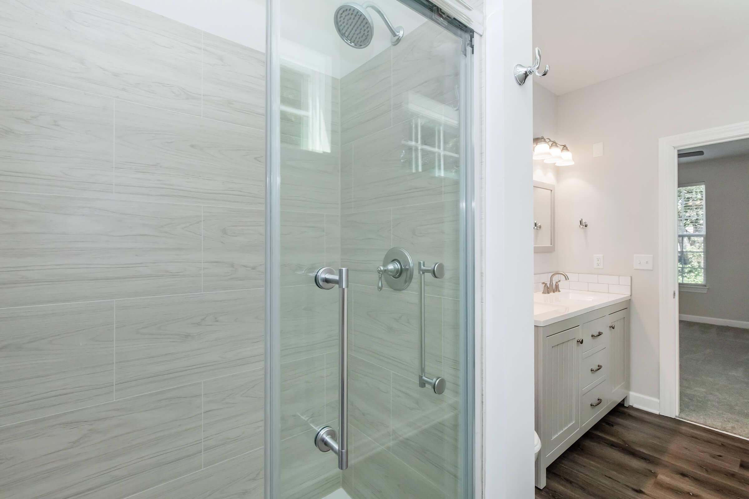 A modern bathroom featuring a glass-enclosed shower with a rainfall showerhead, light-colored wood-style tiles on the walls, and a vanity with a sink. Natural light enters through an adjacent window, illuminating the neutral color scheme and contemporary fixtures.