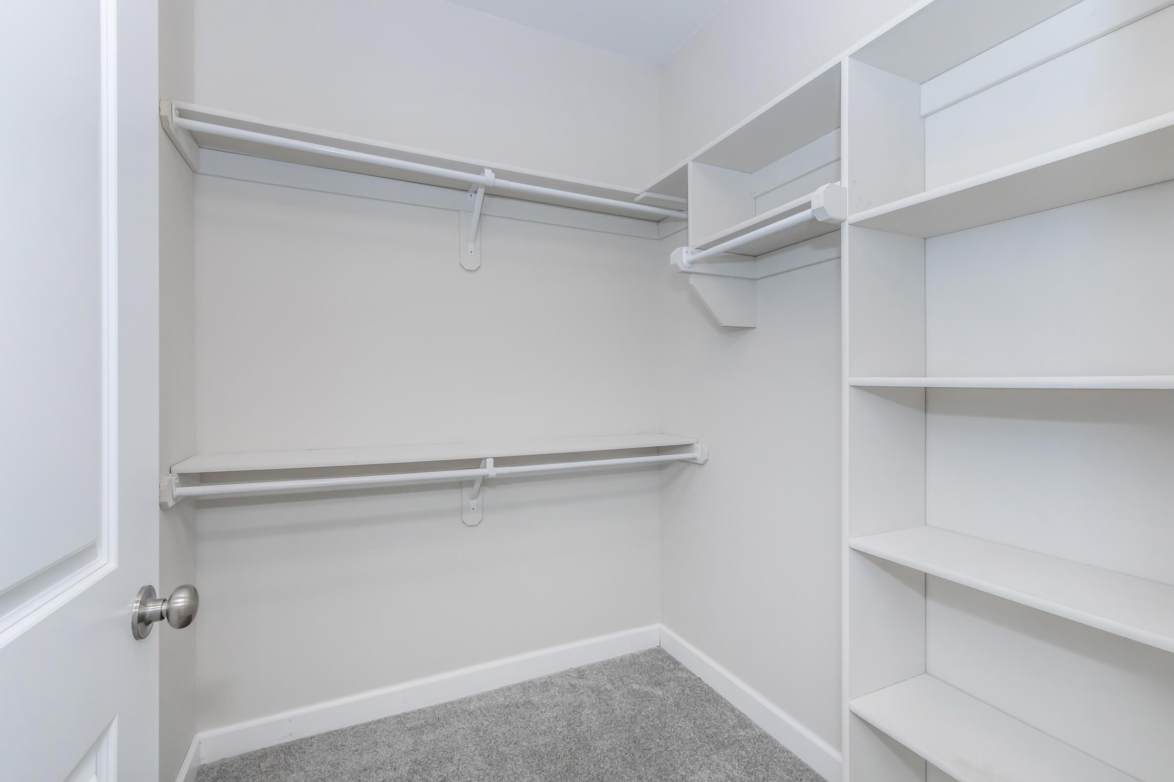 Empty closet with white shelving and hanging rods, featuring a neutral wall color and gray carpet flooring. The space is well-lit and uncluttered, designed for organization and storage.