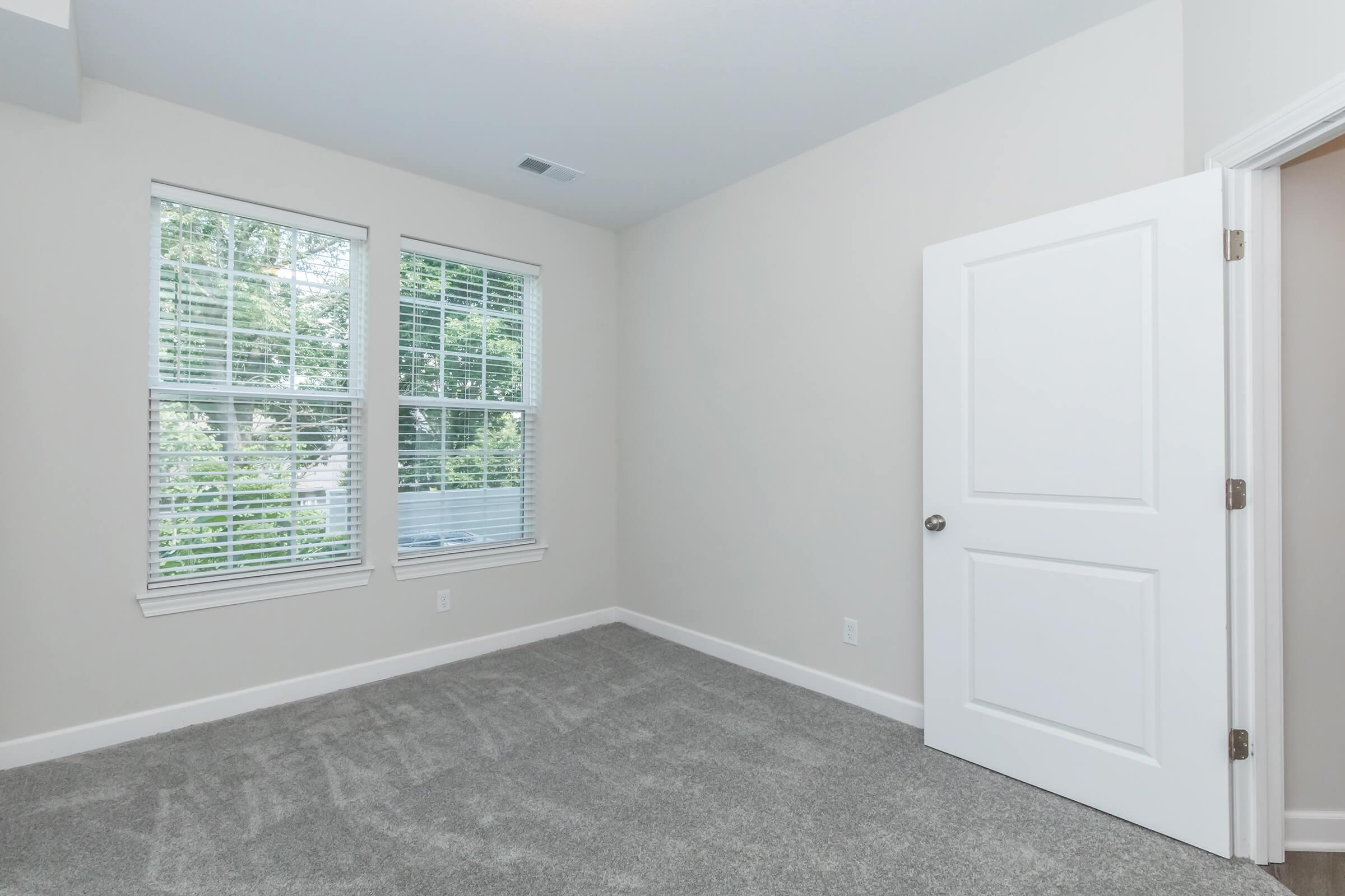 A well-lit, empty bedroom featuring light gray carpet and neutral-colored walls. Two windows with white blinds allow natural light to filter in. A white door is partially open, revealing another doorway or hallway. The space appears clean and ready for personalization.
