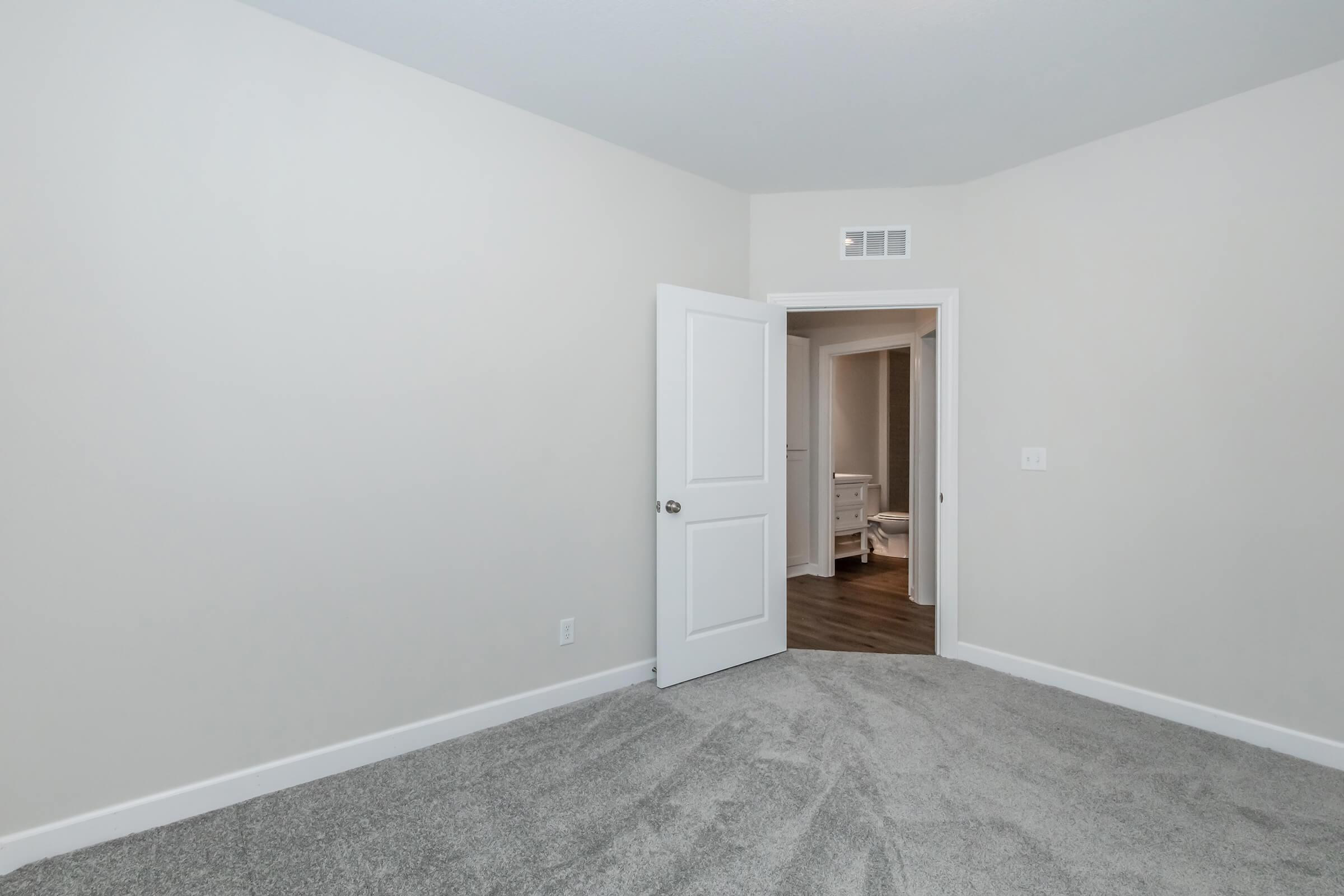 Empty room with light gray carpet and neutral-colored walls. A white door is slightly ajar, leading to another room in the background. The space appears clean and modern, suitable for various uses such as a bedroom or office. Natural light might be coming from an unseen window.