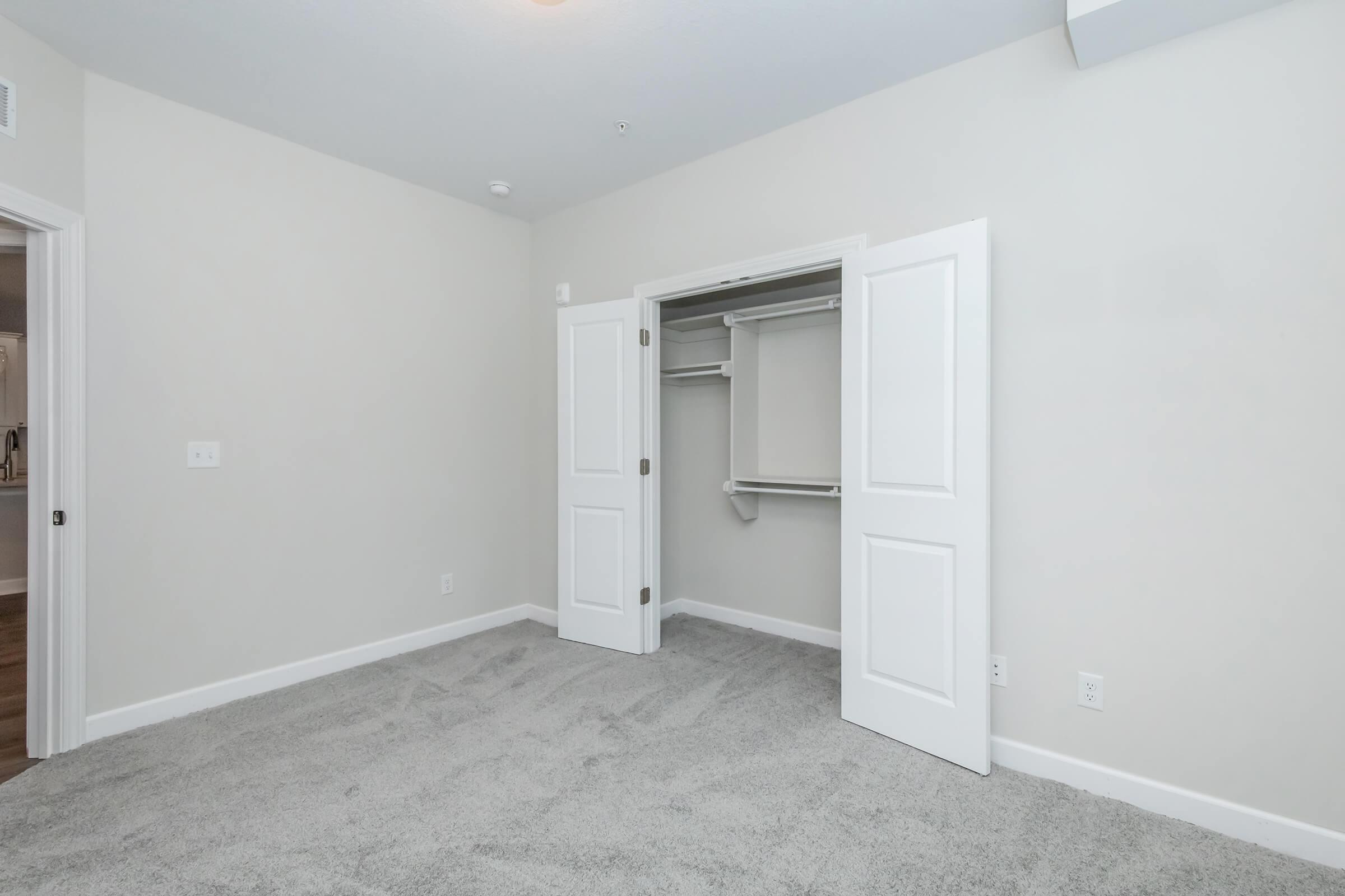 A minimalist bedroom with light gray carpet and neutral-colored walls. There is a small closet with double doors on the left, showing a simple shelving unit inside. The room has no furniture, creating a spacious and empty feel. Natural light may be coming from an unseen window.