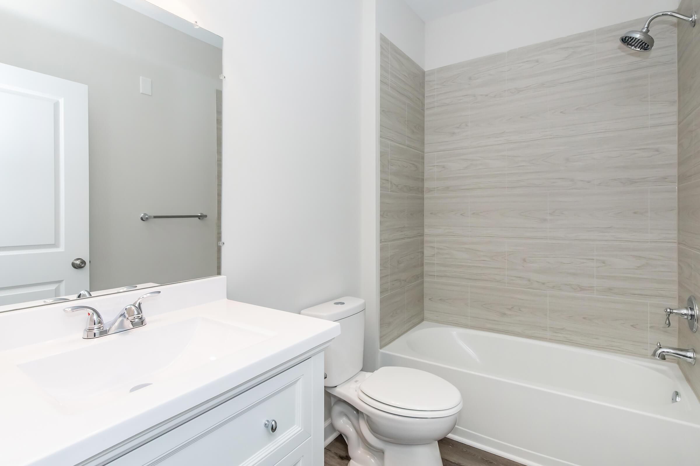 A bright, modern bathroom featuring a white tub and shower with a sleek chrome showerhead. The vanity has a white countertop with two faucets, and there is a large mirror above. Light-colored, textured wall tiles complement the space. A door leads to another room, enhancing the airy feel.