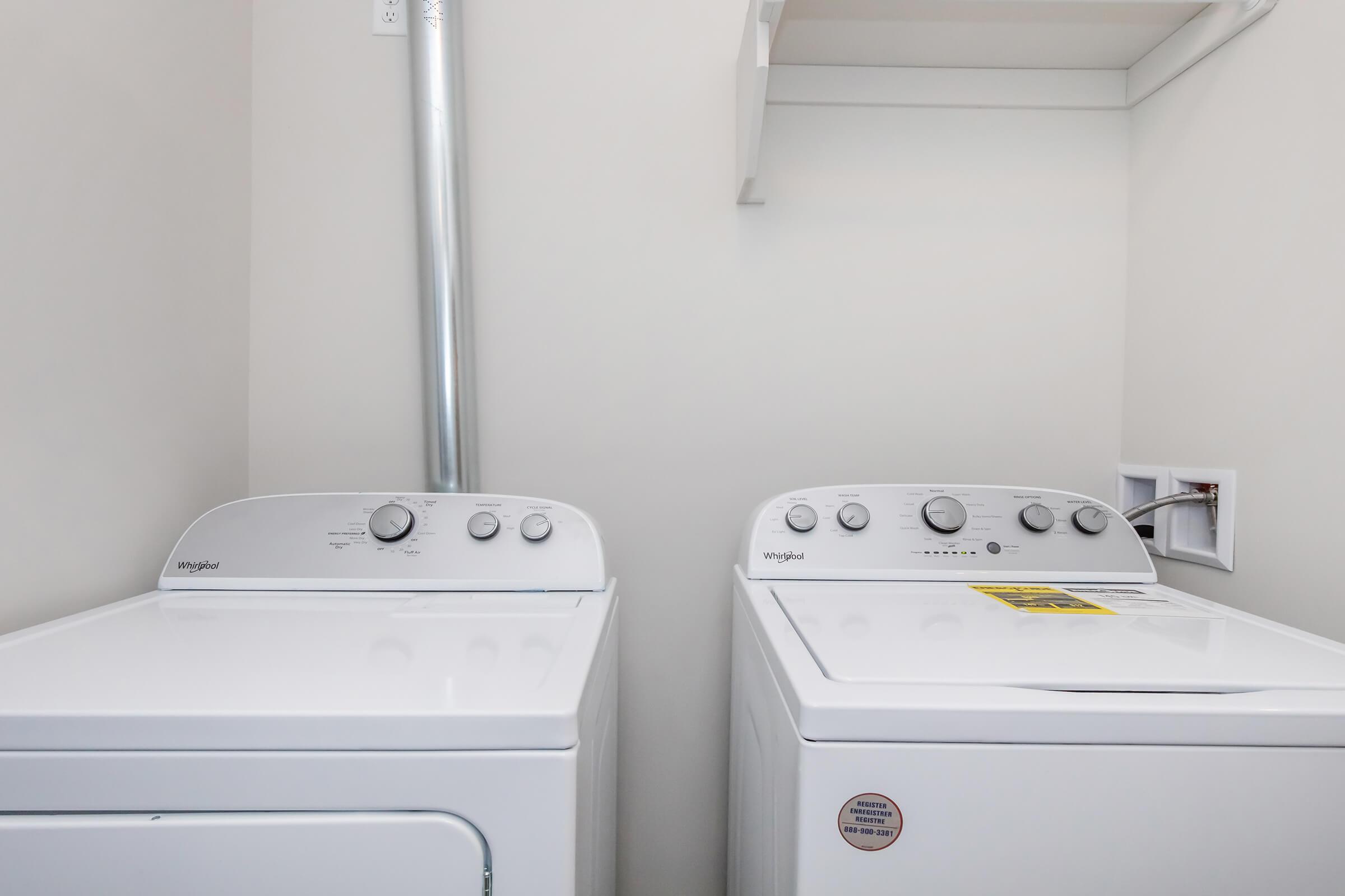 Two white washing machines side by side in a laundry room. The left machine is a front-loader, while the right machine is a top-loader. Both have control dials on the front, and there is a wall shelf above them. The setting is bright and clean with a neutral wall color.
