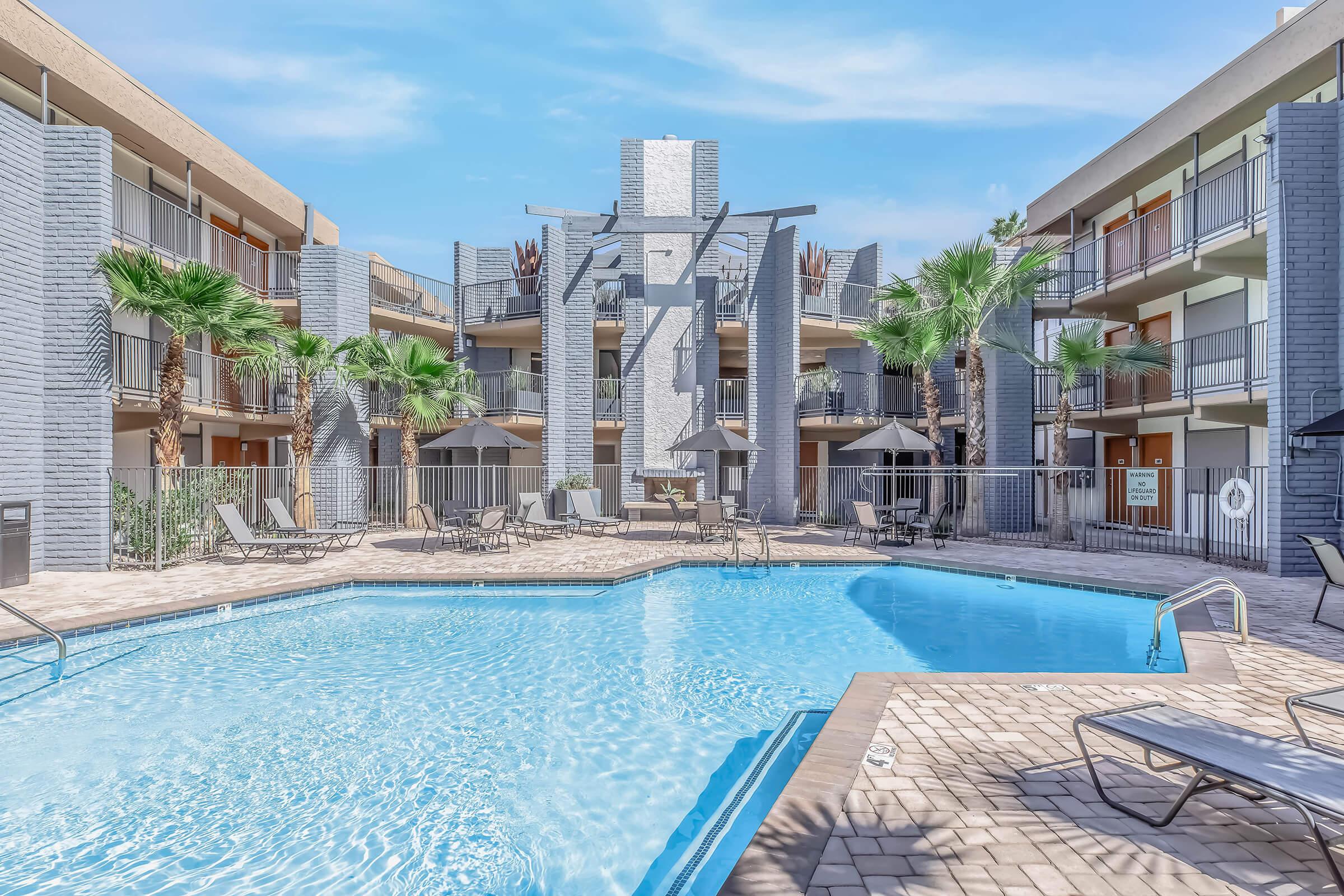 A spacious pool area surrounded by modern apartment buildings, featuring lounge chairs and palm trees. The pool is clear and inviting, with a unique sculptural feature in the center and shaded seating areas under umbrellas. Bright, sunny skies complete the pleasant atmosphere.