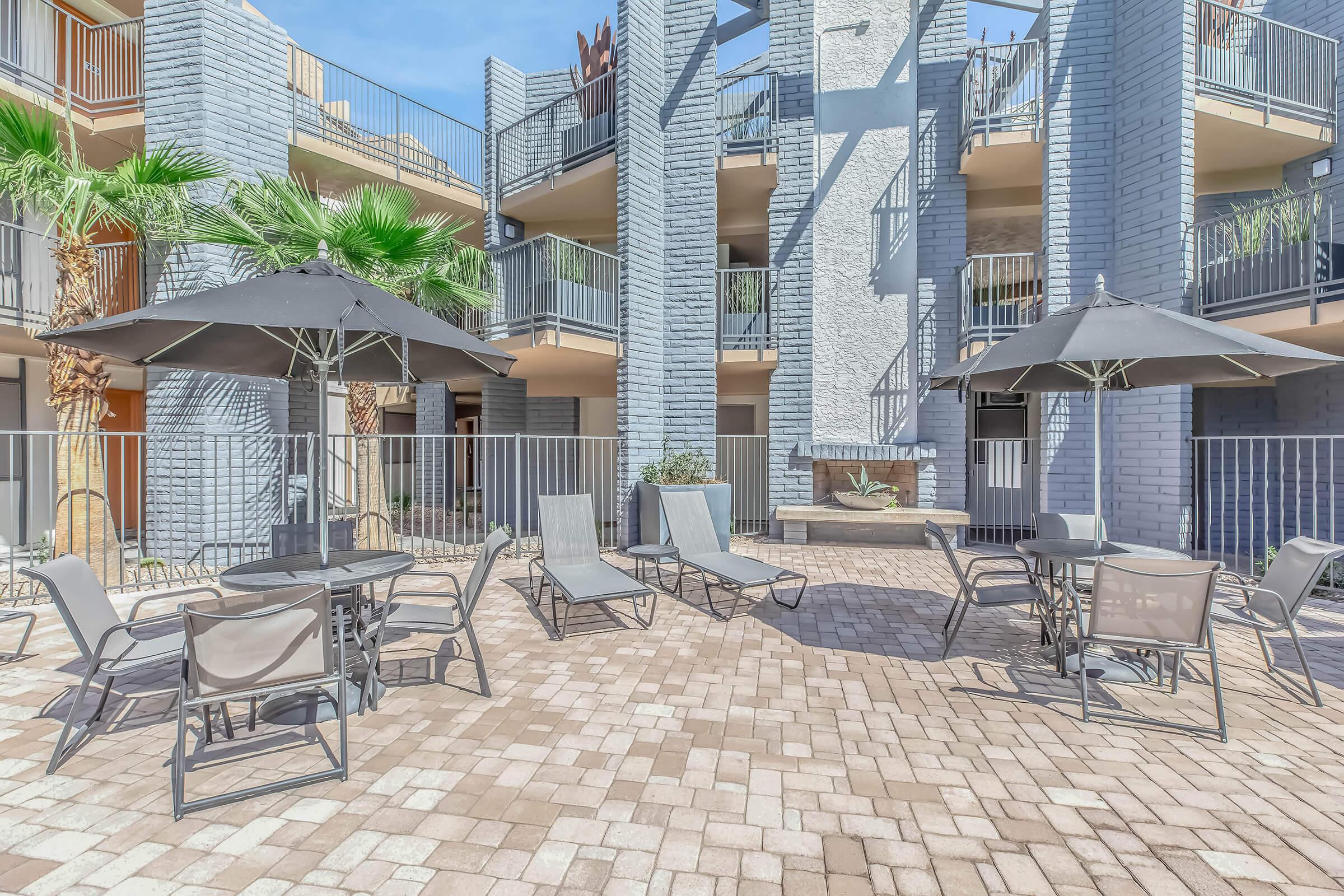 A tranquil outdoor area with lounge chairs and tables under large umbrellas, surrounded by palm trees and modern apartment buildings. The space features a clean, paved surface, ideal for relaxation and socializing. Bright blue sky enhances the inviting atmosphere.