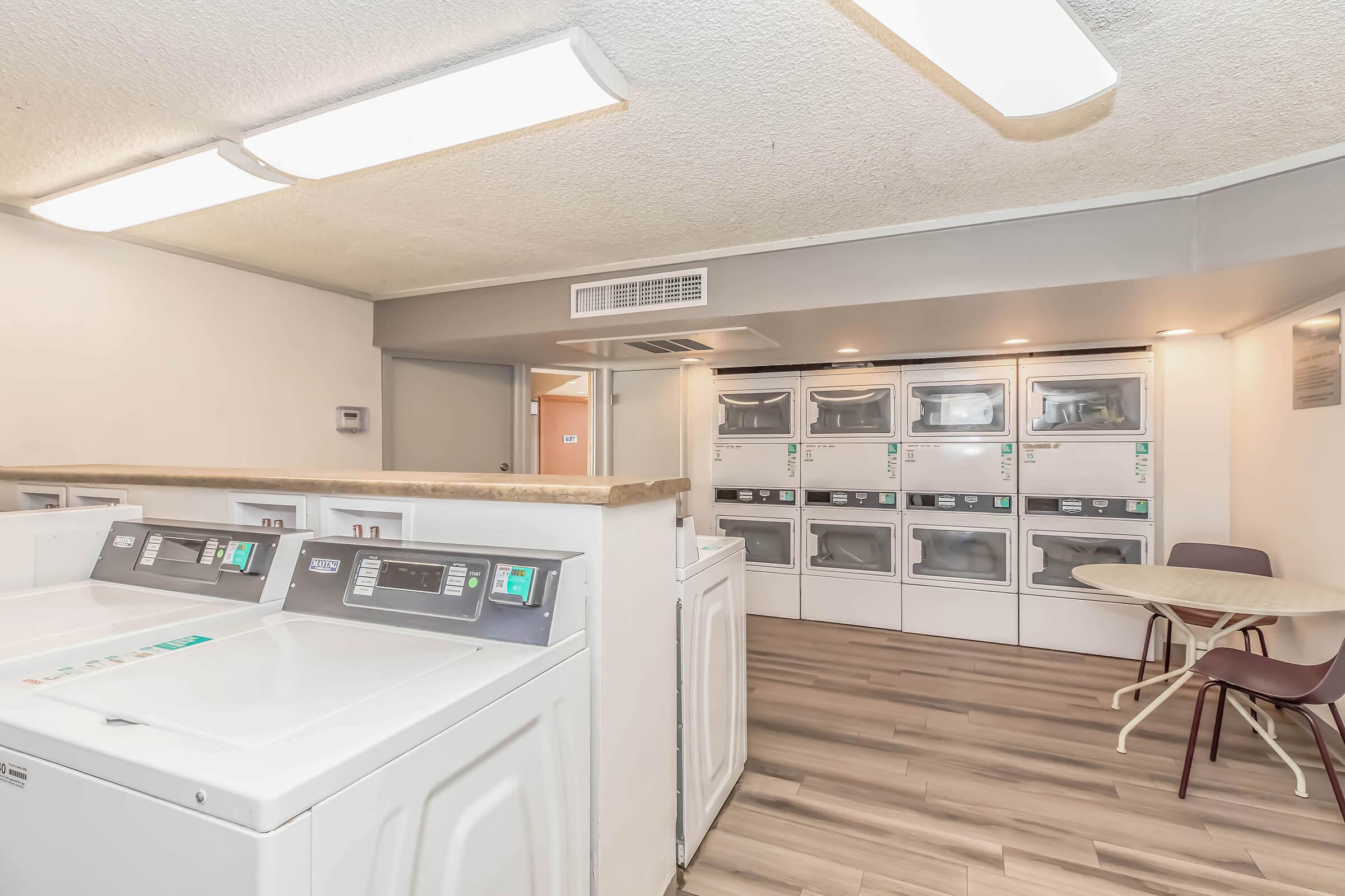 Bright and clean laundry room featuring several commercial washing machines and dryers, a table with a chair, and a well-lit atmosphere with ceiling lights. The floors are wooden, and the walls are painted in neutral colors, creating a welcoming space for laundry activities.