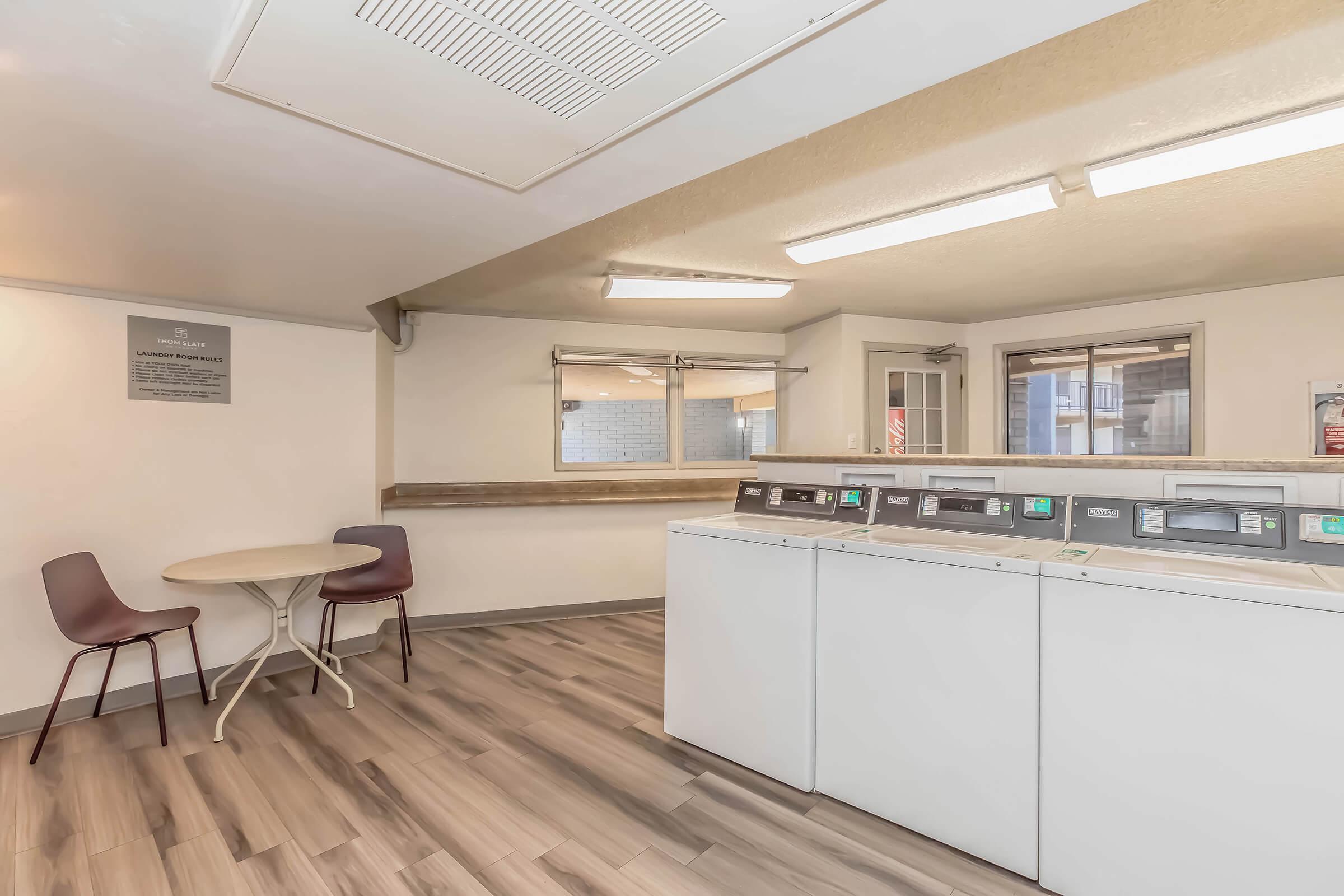 A laundry room with several washing machines and dryers, featuring a small round table with two chairs nearby. The space has white walls and a wood-like floor, with bright overhead lighting. A sign is mounted on the wall, providing information about the laundry facilities.