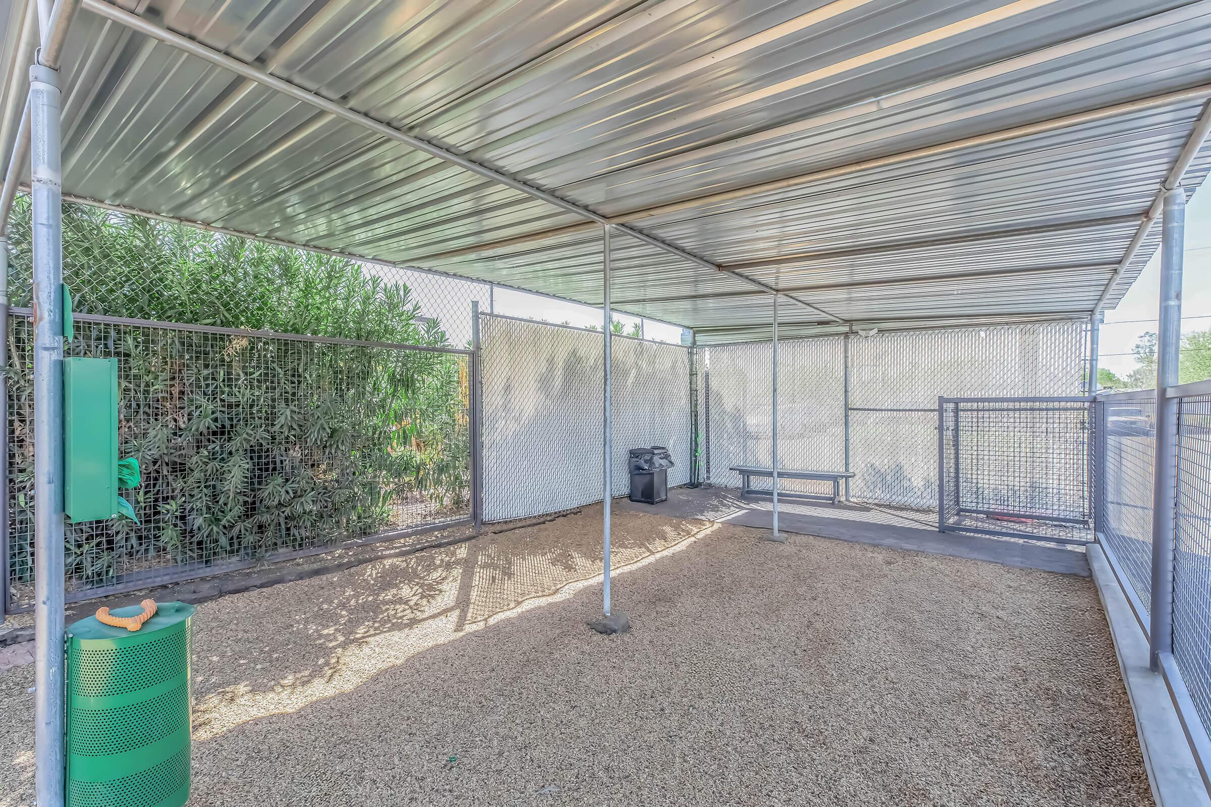A spacious outdoor enclosure with a gravel floor, surrounded by chain-link fencing and covered by a metal roof. There are green waste bins, a bench, and nearby shrubs, providing a shaded area suitable for pets or outdoor activities.
