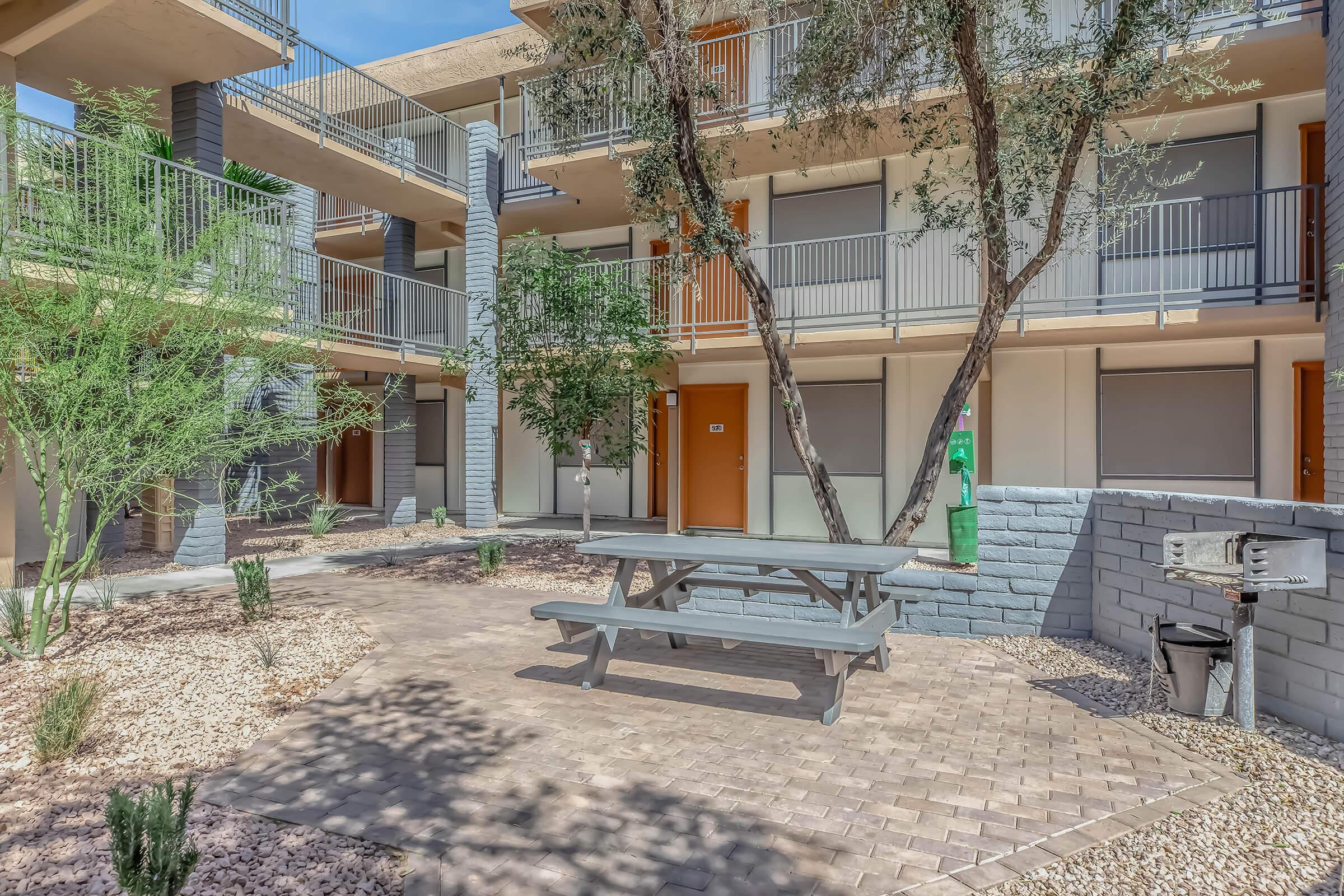 A tranquil courtyard featuring a picnic table surrounded by trees and shrubs. The area has access to multiple apartment balconies with wooden doors and railings. A barbeque grill is positioned nearby, offering a place for outdoor cooking and dining amidst a well-maintained landscape.