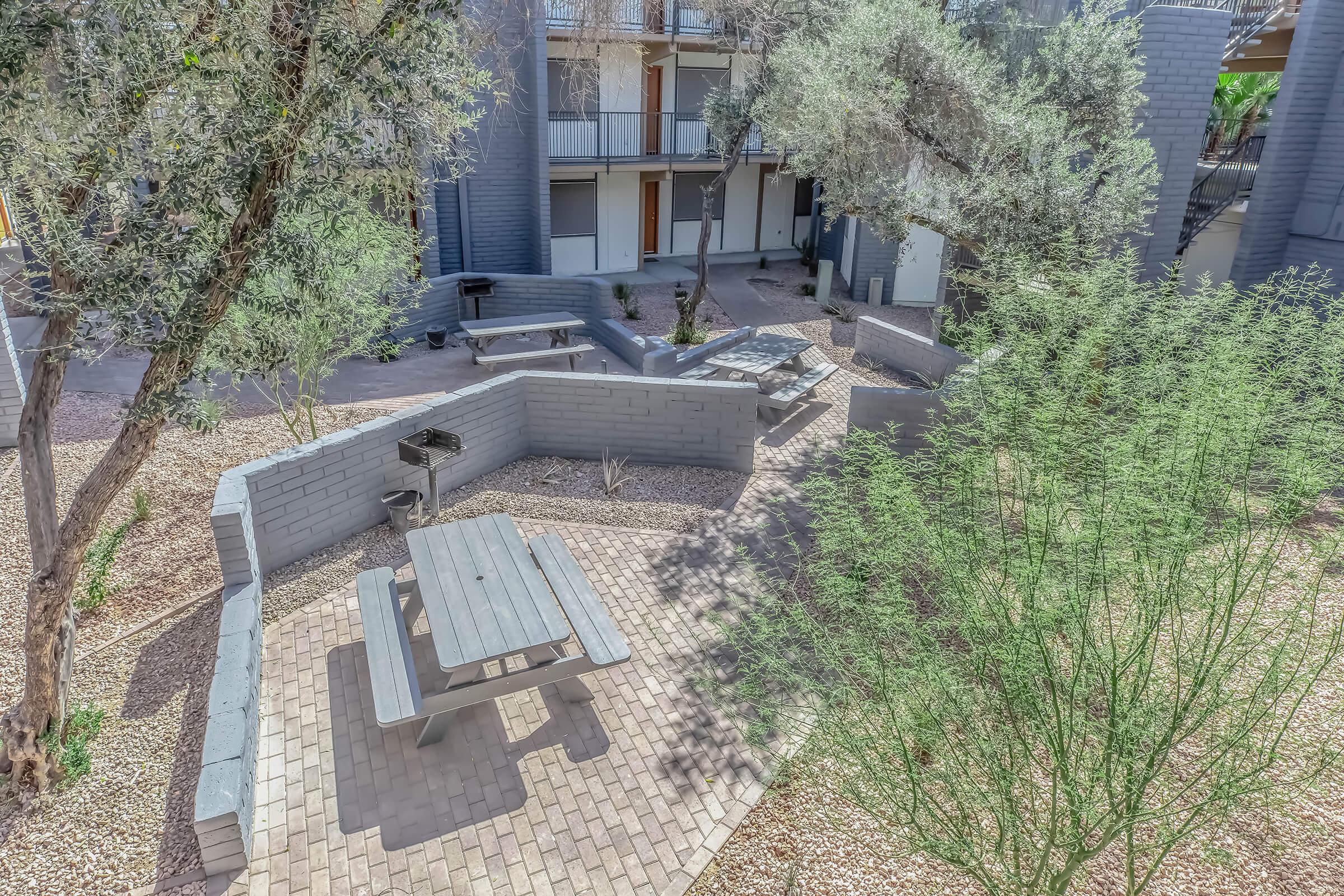 A courtyard area featuring grey brick pathways, picnic tables, and barbecue grills surrounded by greenery and trees. The space is designed for outdoor gatherings, with a modern building visible in the background, showcasing multiple balcony entrances.