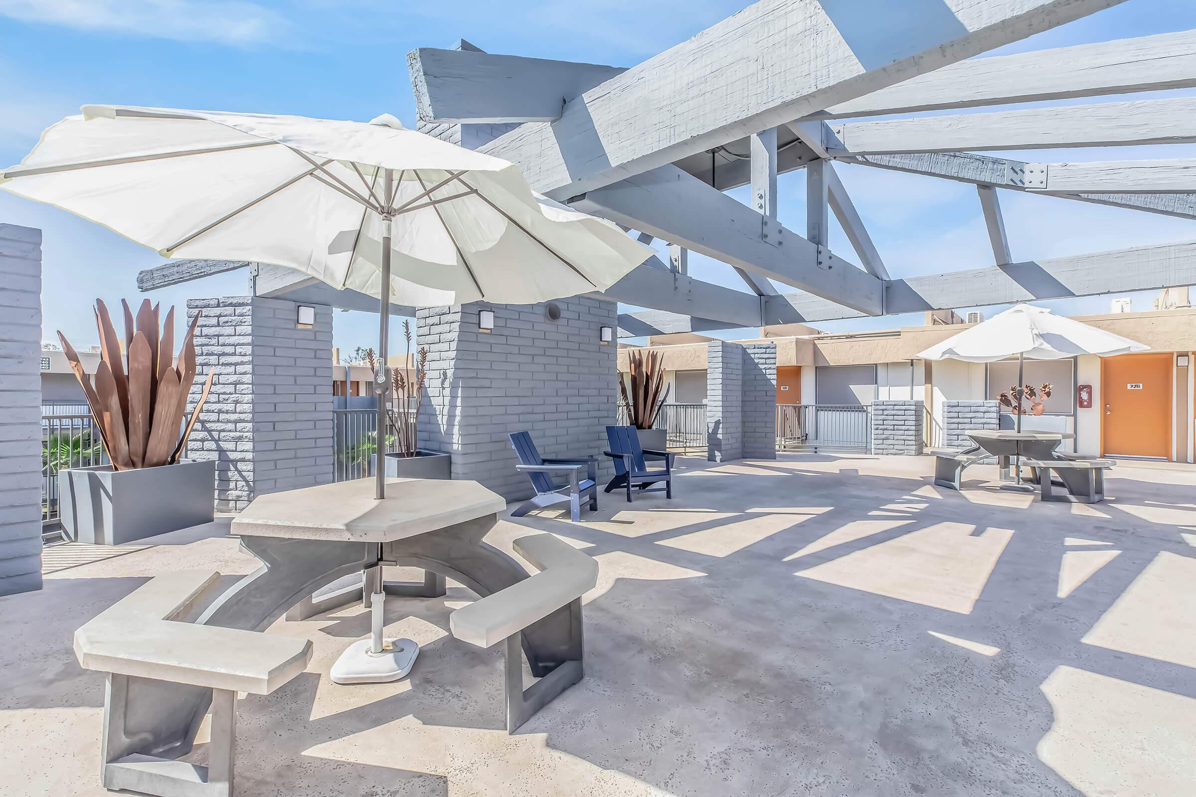 A rooftop terrace featuring modern outdoor furniture, including a circular concrete table with attached benches, several blue lounge chairs, and large white umbrellas. The area is adorned with architectural beams and decorative plants, providing a spacious and inviting atmosphere for relaxation.