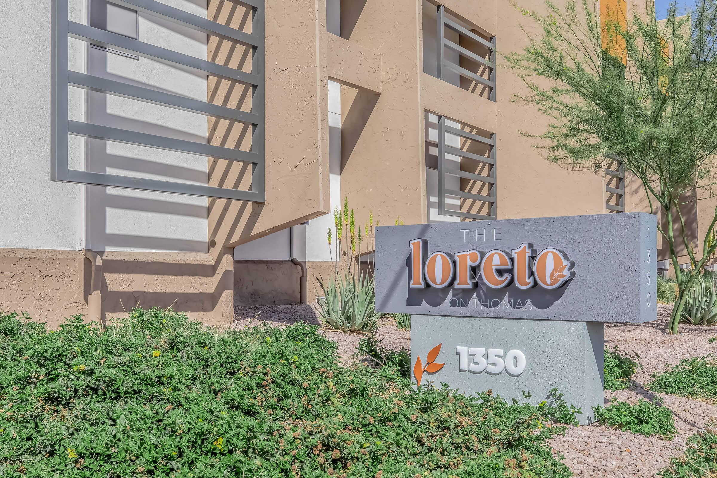 A sign for "The Loreto" with the address "1350" displayed prominently. The sign is situated in front of a building with desert landscaping, including green shrubs and cacti, against a neutral-colored wall.