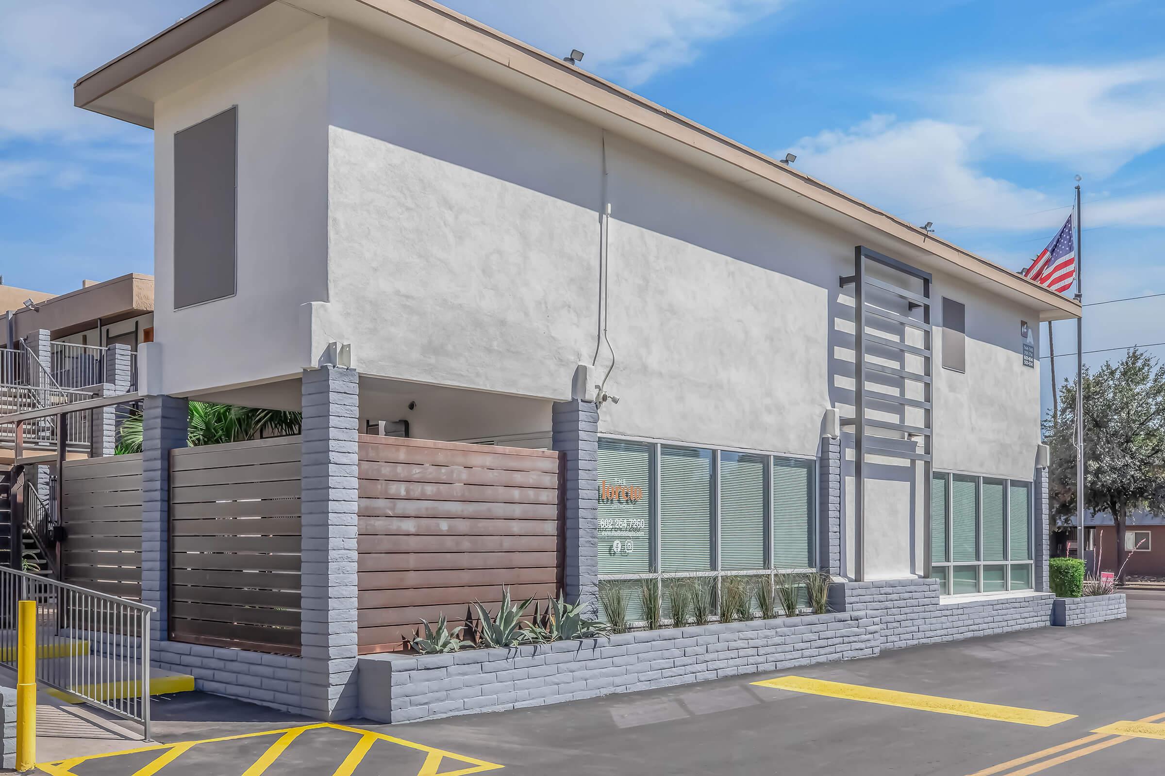 A modern, two-story building with a clean, minimalist design. The exterior features a light-colored facade, large windows, and wooden accents. A U.S. flag is visible on the side. The surrounding area includes pavement, low-maintenance landscaping, and a clear blue sky.