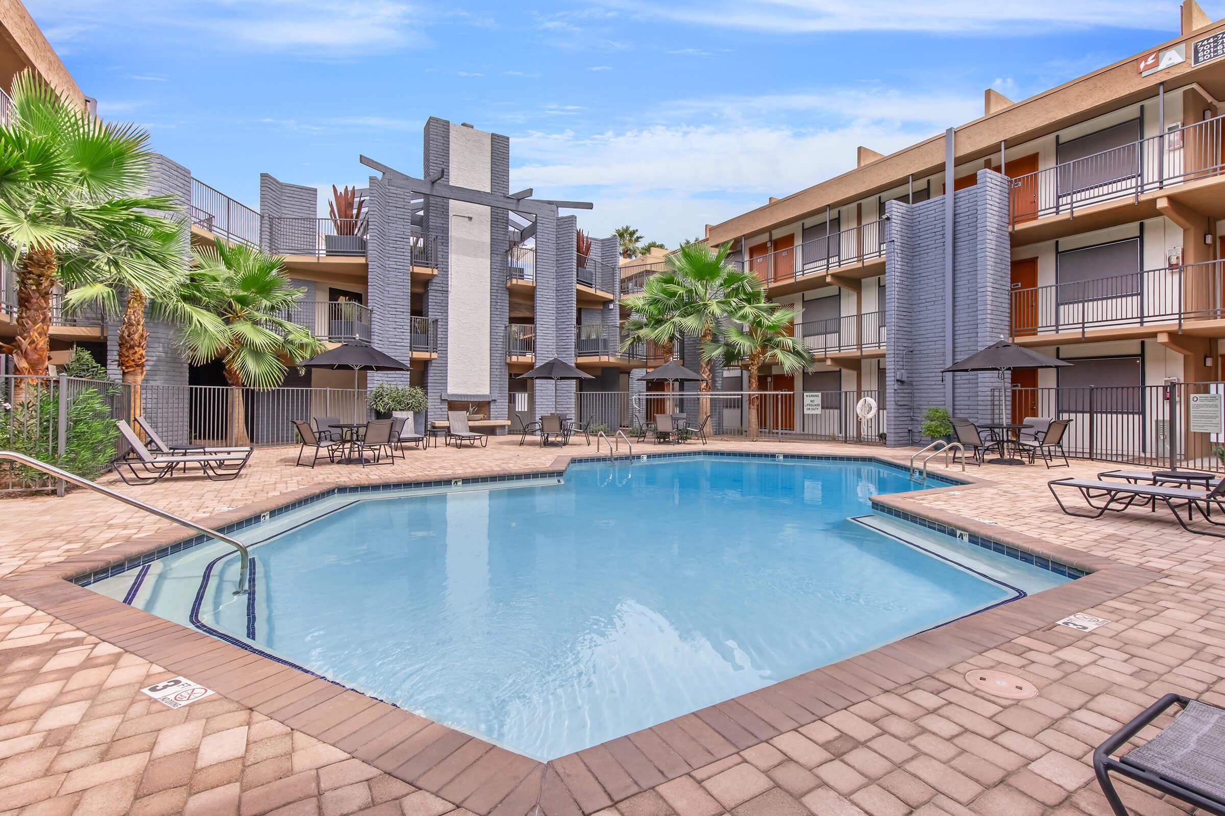 A sunny pool area surrounded by palm trees and lounge chairs, with a large swimming pool in the center. The pool is framed by two multilevel buildings featuring balconies. The setting is inviting and ideal for relaxation, with a clear blue sky overhead.