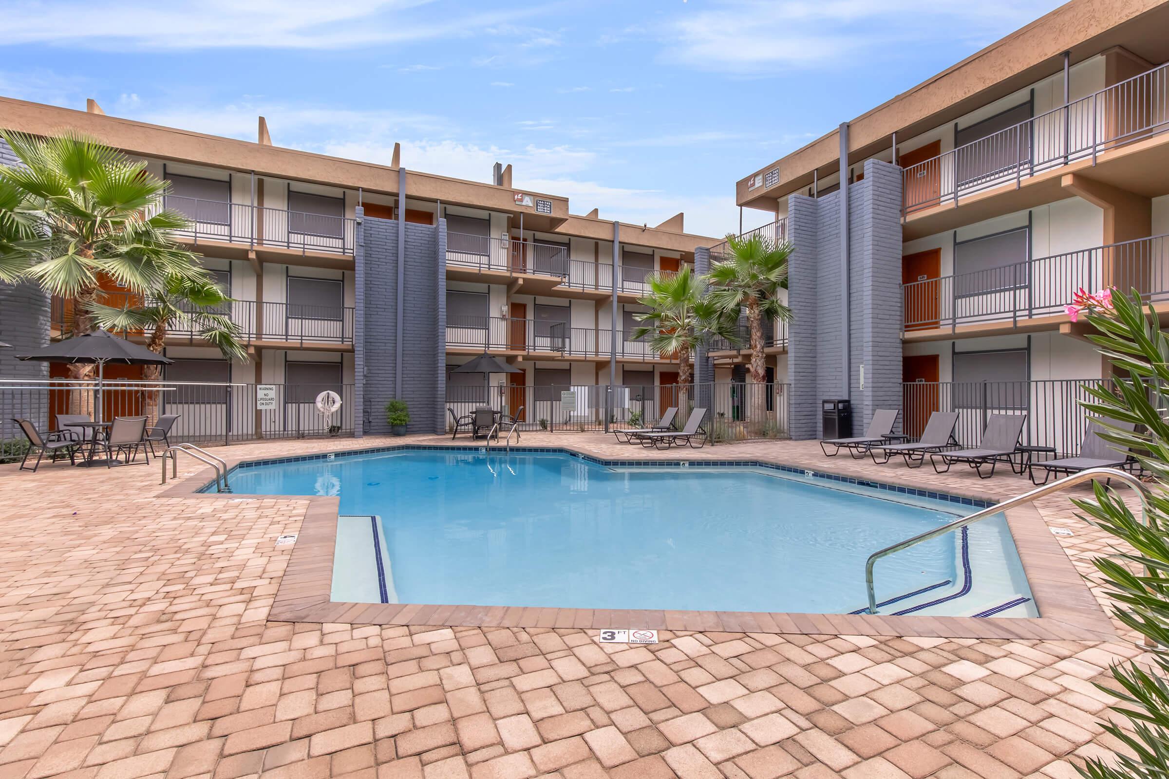 A clean, inviting swimming pool surrounded by lounge chairs and palm trees, situated in a courtyard of a multi-story building with balconies. The setting features a modern design, with clear blue skies reflecting above the pool, creating a relaxing atmosphere.