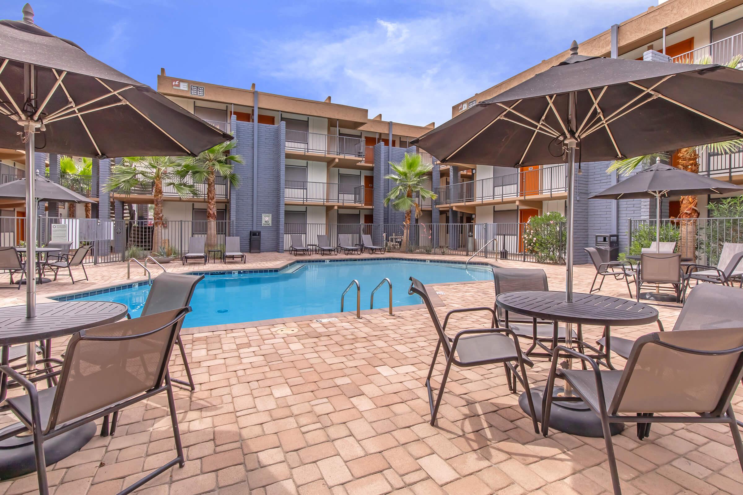 A view of a landscaped courtyard featuring a swimming pool surrounded by palm trees, with several patio tables and umbrellas. The building's balconies overlook the pool area, creating a relaxing outdoor atmosphere suitable for leisure and socializing. Clear blue sky complements the scene.