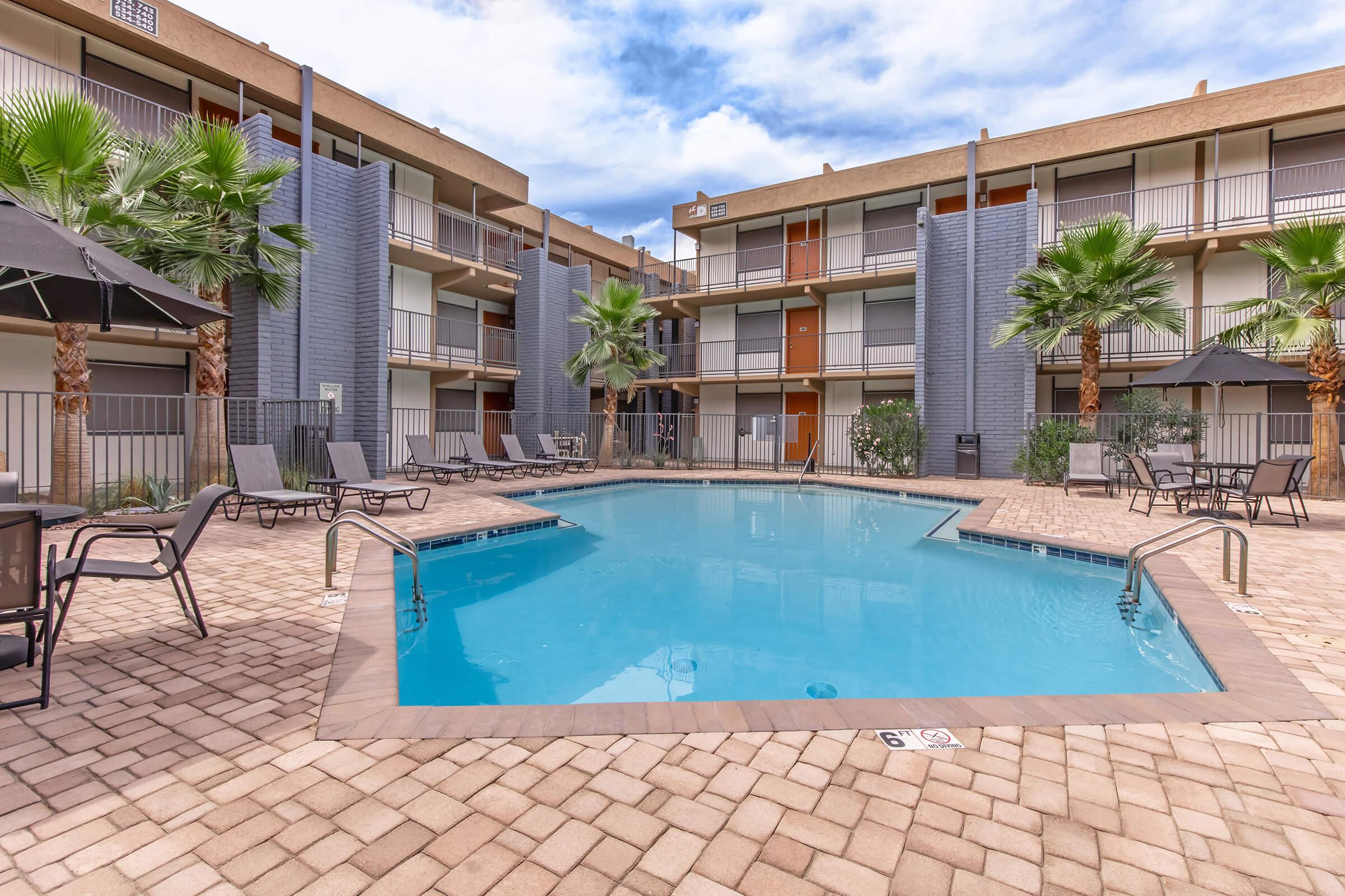 A view of a swimming pool surrounded by palm trees and lounge chairs. The pool area is bordered by two-story apartment buildings featuring balconies. The sky is partly cloudy, creating a pleasant atmosphere for relaxation.