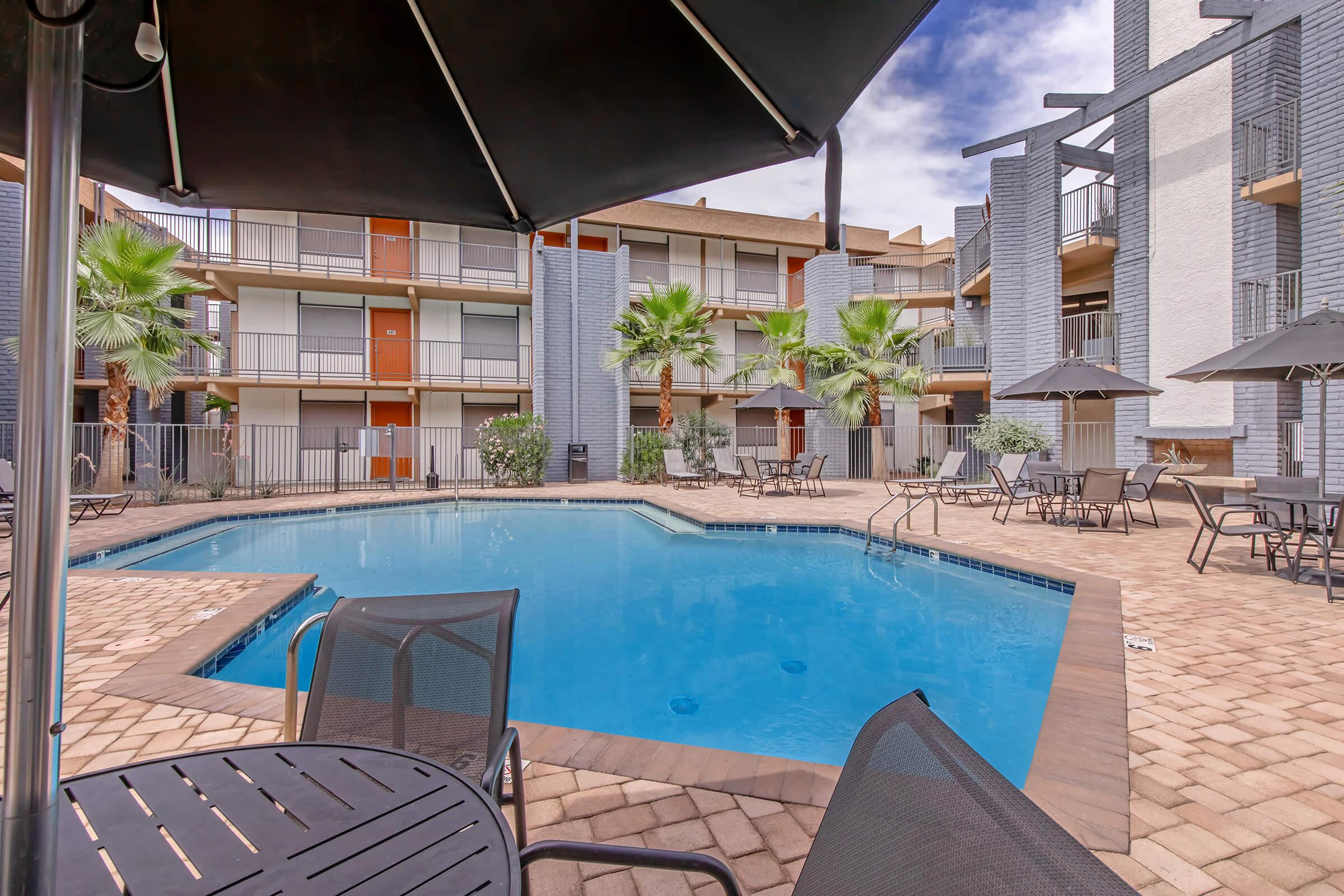 A view of a residential apartment complex featuring a swimming pool surrounded by palm trees and sun umbrellas. Patio seating is visible, with two buildings in the background showing balconies. The sky is partly cloudy, and the setting conveys a relaxed, inviting atmosphere.