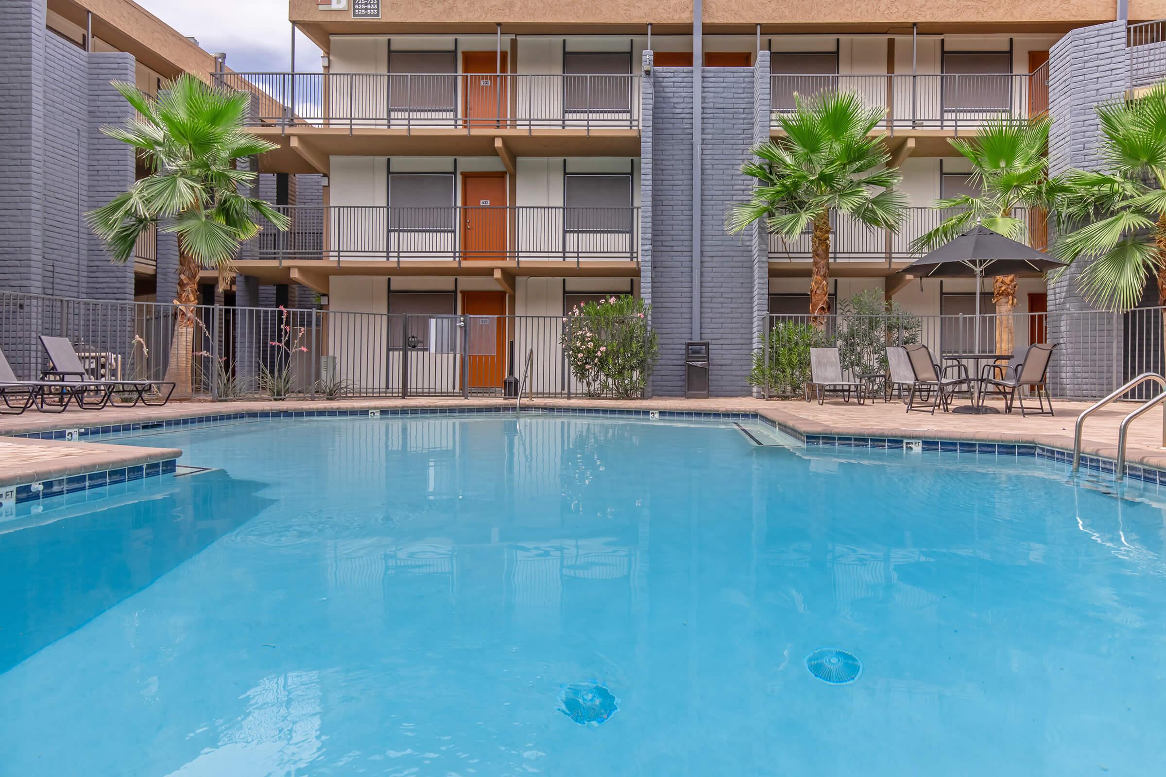 A clear swimming pool surrounded by a wooden deck, with palm trees on either side. In the background, there are two-story buildings featuring balconies and doors. The atmosphere is inviting and tropical, perfect for relaxation and leisure.