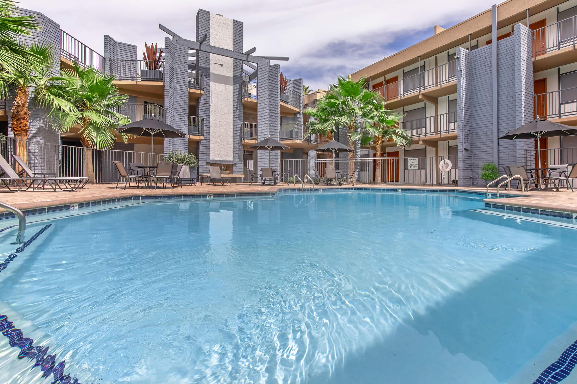A swimming pool surrounded by a modern courtyard featuring palm trees, lounge chairs, and umbrellas. The pool area is flanked by multi-story buildings with balconies. The sky is partly cloudy, creating a serene atmosphere ideal for relaxation.