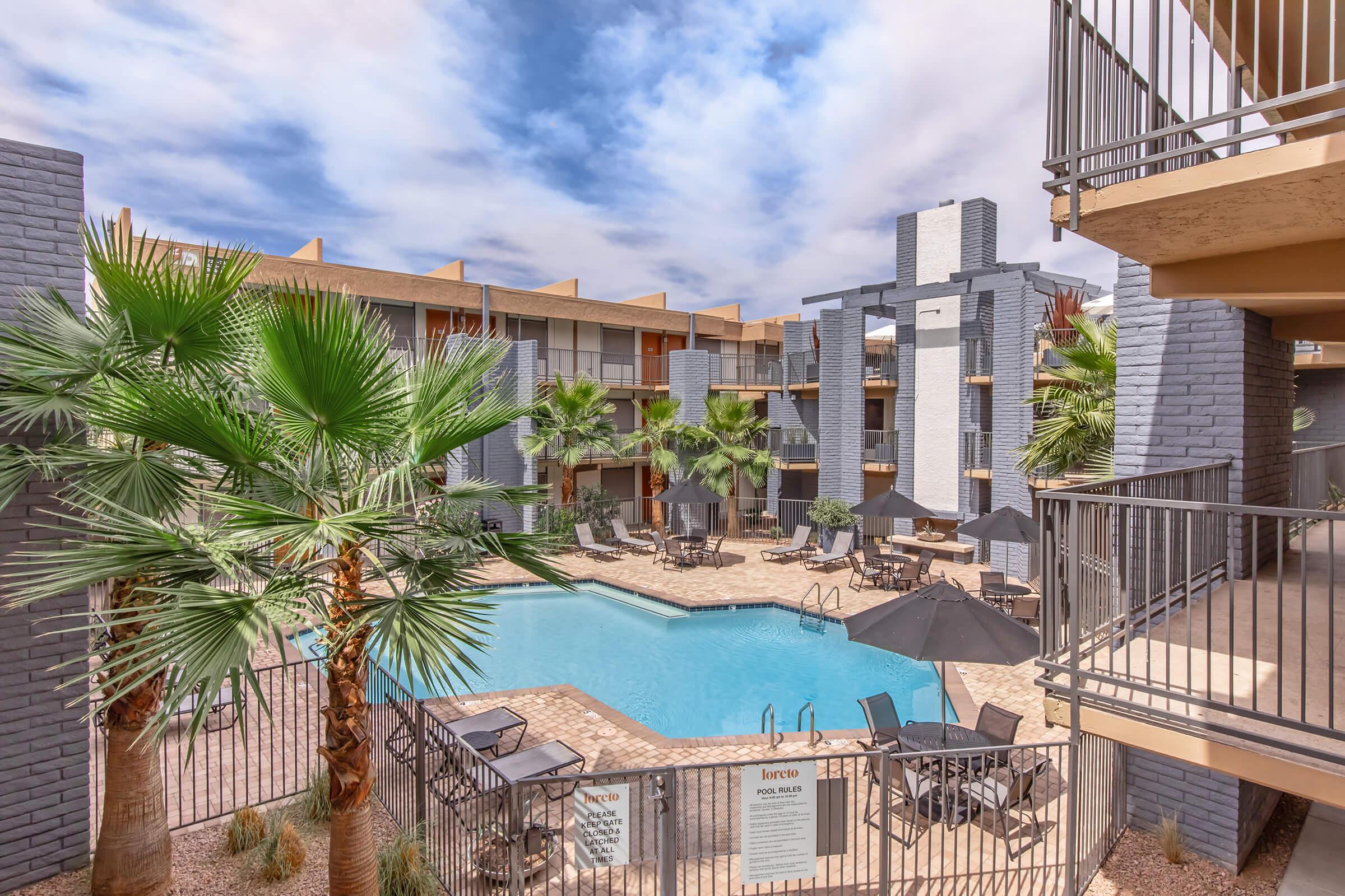 A vibrant outdoor swimming pool surrounded by palm trees and lounge chairs, located in a modern apartment complex. The architecture features contemporary designs with gray brick walls. The sky above is partially cloudy, enhancing the inviting atmosphere of the pool area.