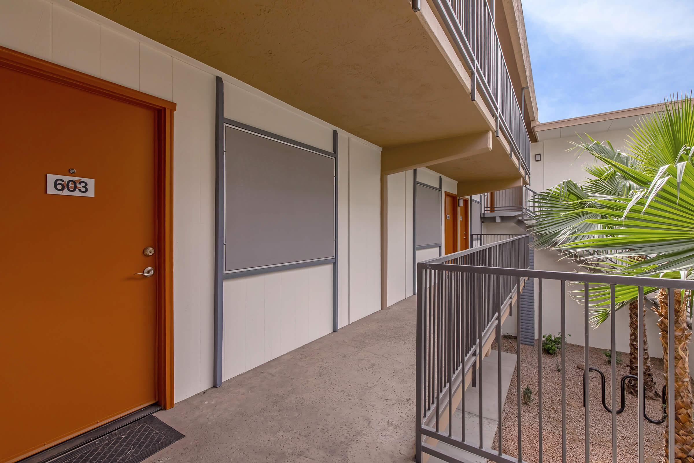 A well-maintained apartment balcony with two doors numbered 603 and a clear view of a palm tree. The exterior features light-colored walls, orange doors, and gray railings, with a concrete walkway leading to the entrance.