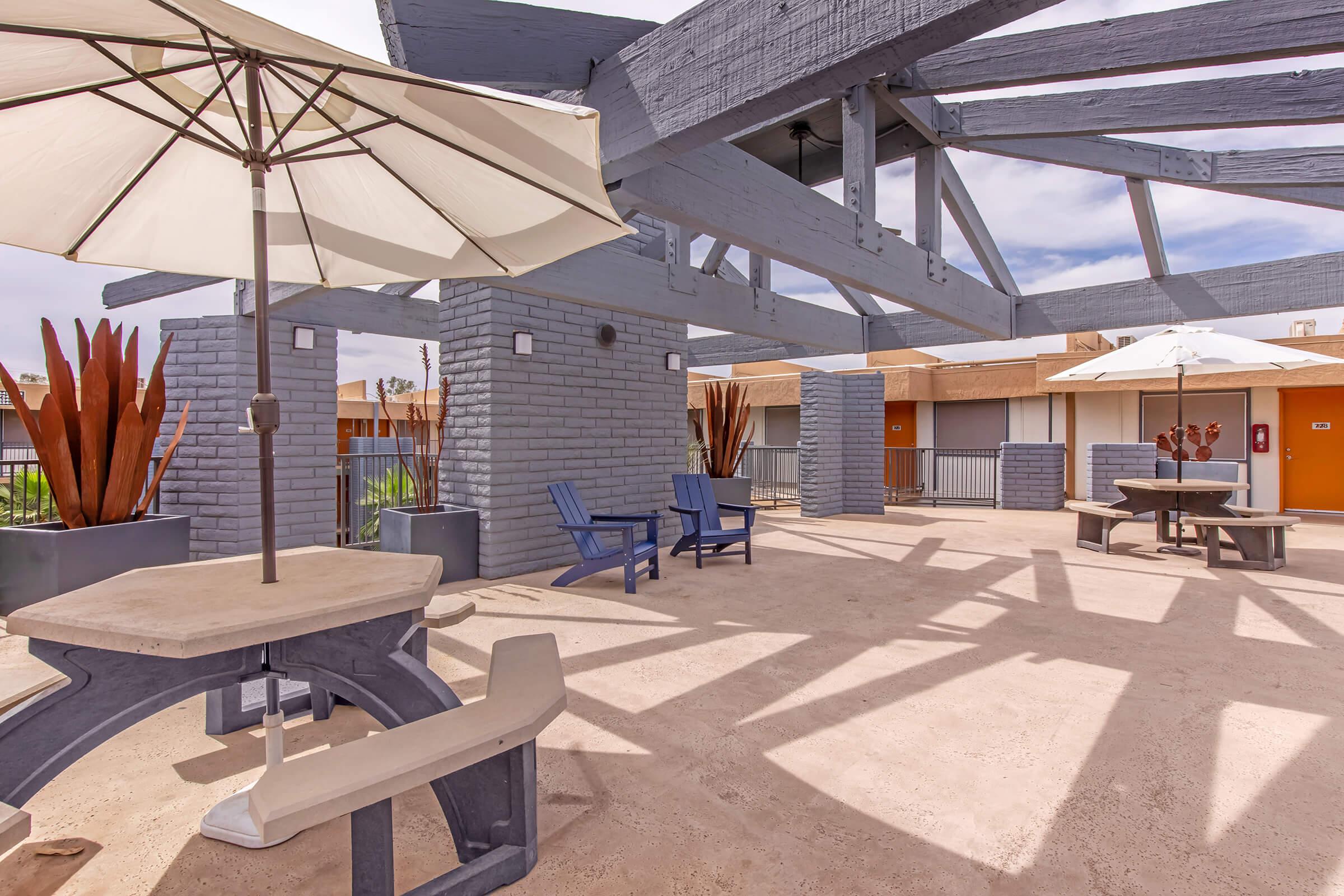 An outdoor patio area featuring blue wooden chairs and a table, shaded by large umbrellas. The space is surrounded by potted plants and has a modern design with gray brick columns and a wooden overhead structure, creating a relaxing atmosphere.