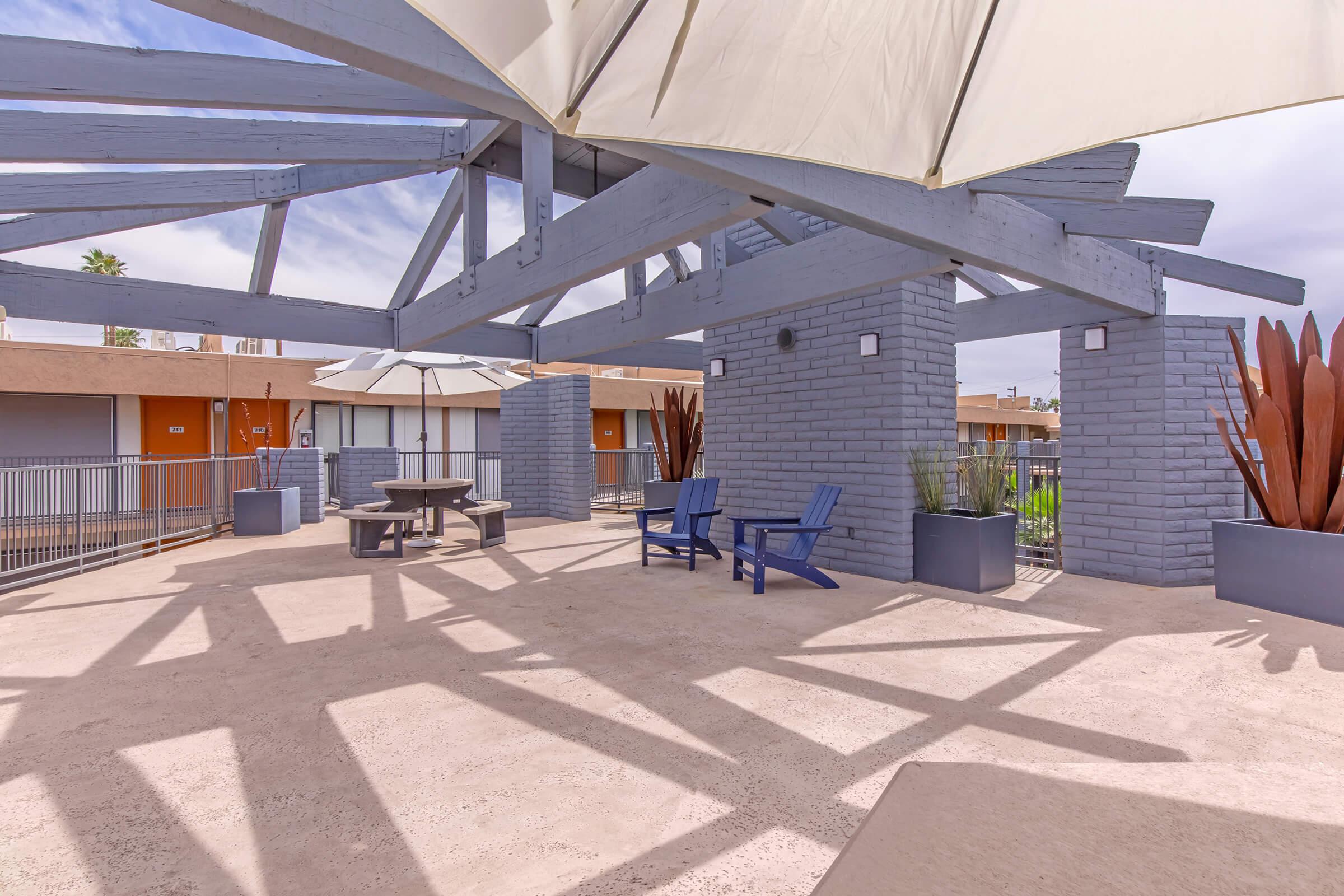 A spacious outdoor area featuring a shaded seating space with blue Adirondack chairs, a round table, and large planters. The structure has a modern architectural design with beams and a canvas cover, providing shade. The background shows a low-rise building with multiple entrances.