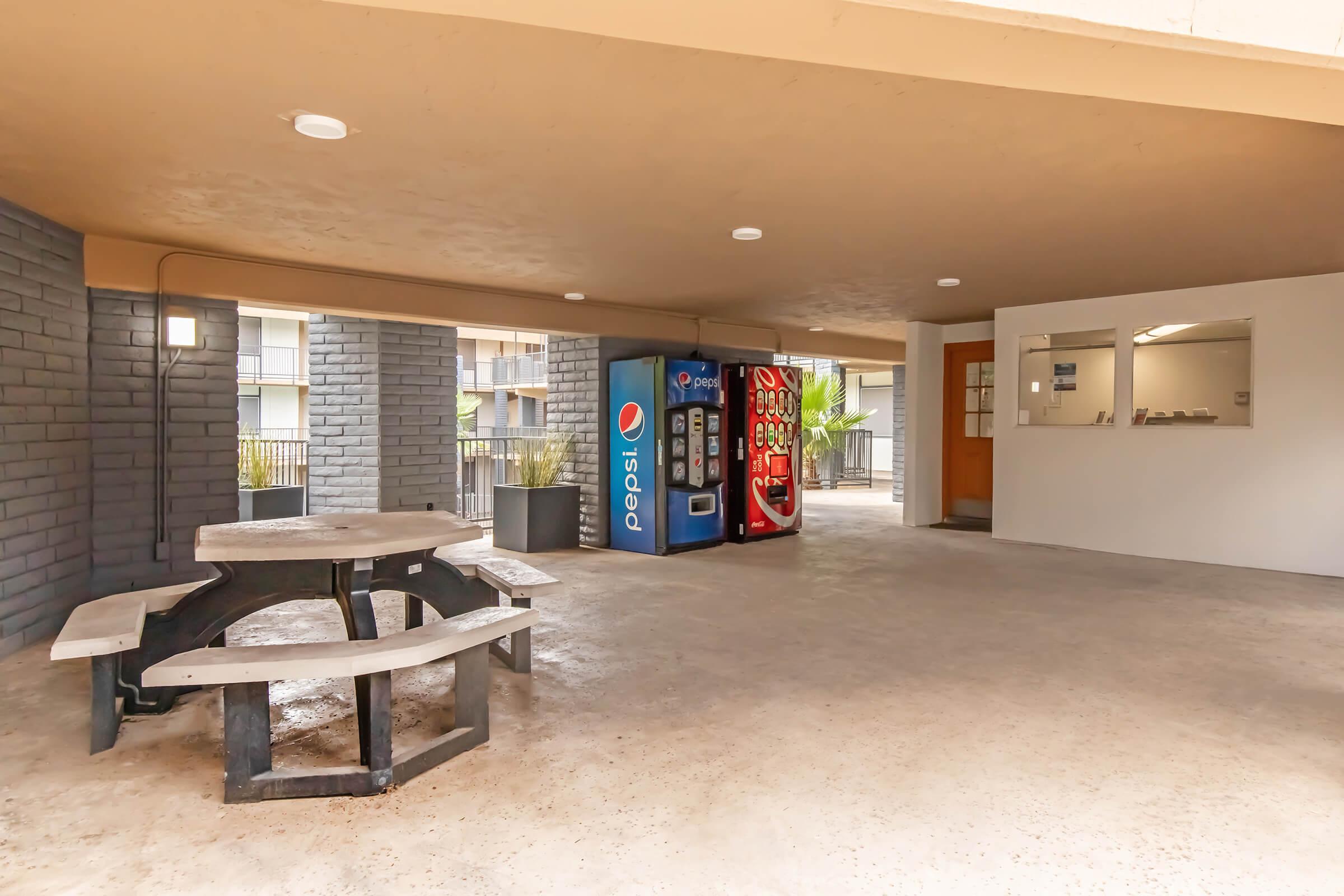 A covered outdoor area features a picnic table, two vending machines displaying Pepsi and snacks, and an office entrance. The space is well-lit with gray brick walls and a concrete floor, providing a casual gathering spot for residents.