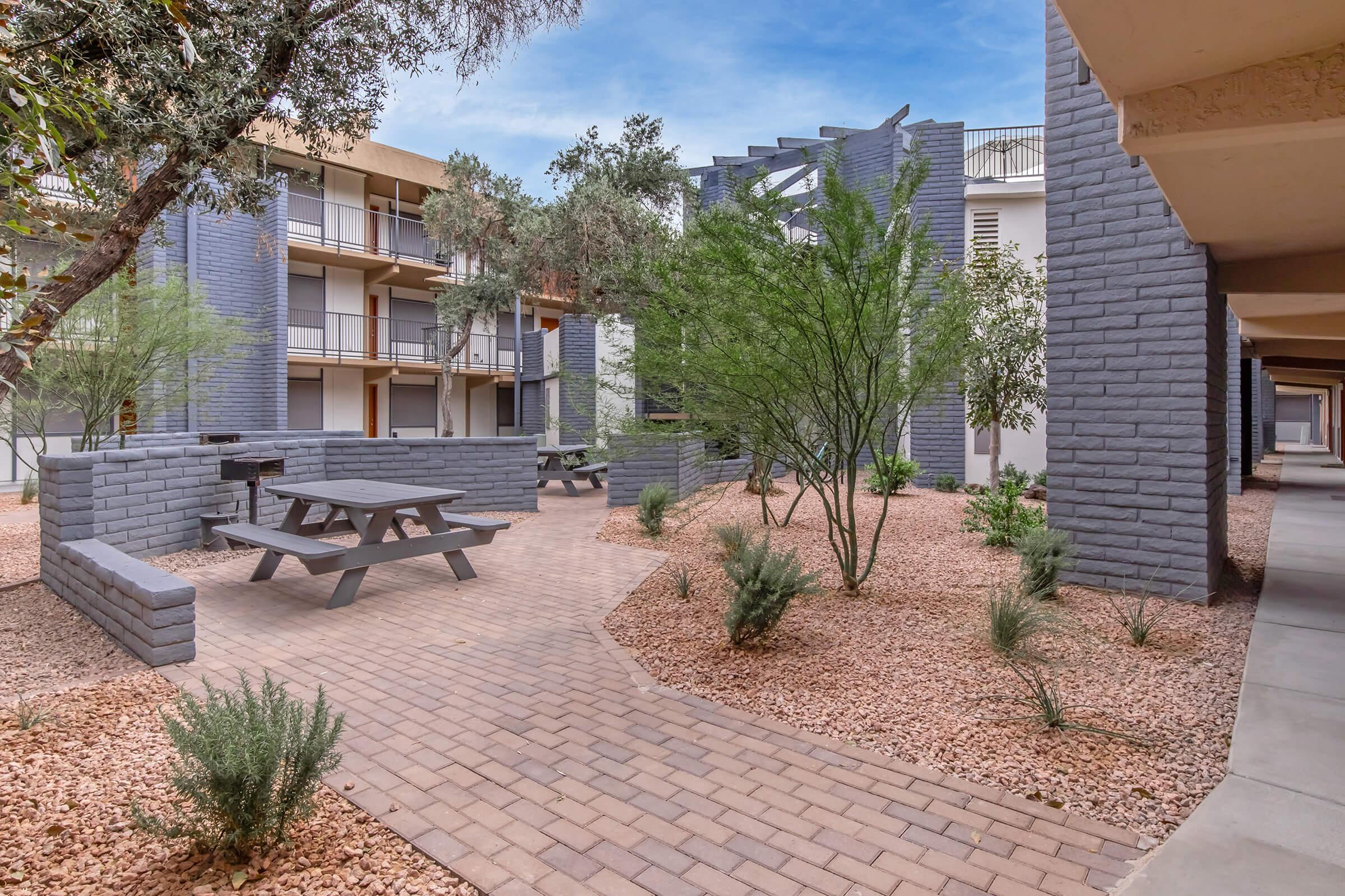 A landscaped courtyard featuring brick pathways, greenery, and picnic tables. Surrounding buildings have a modern design with a mix of gray and beige colors. The scene is bright and inviting, ideal for outdoor gatherings.