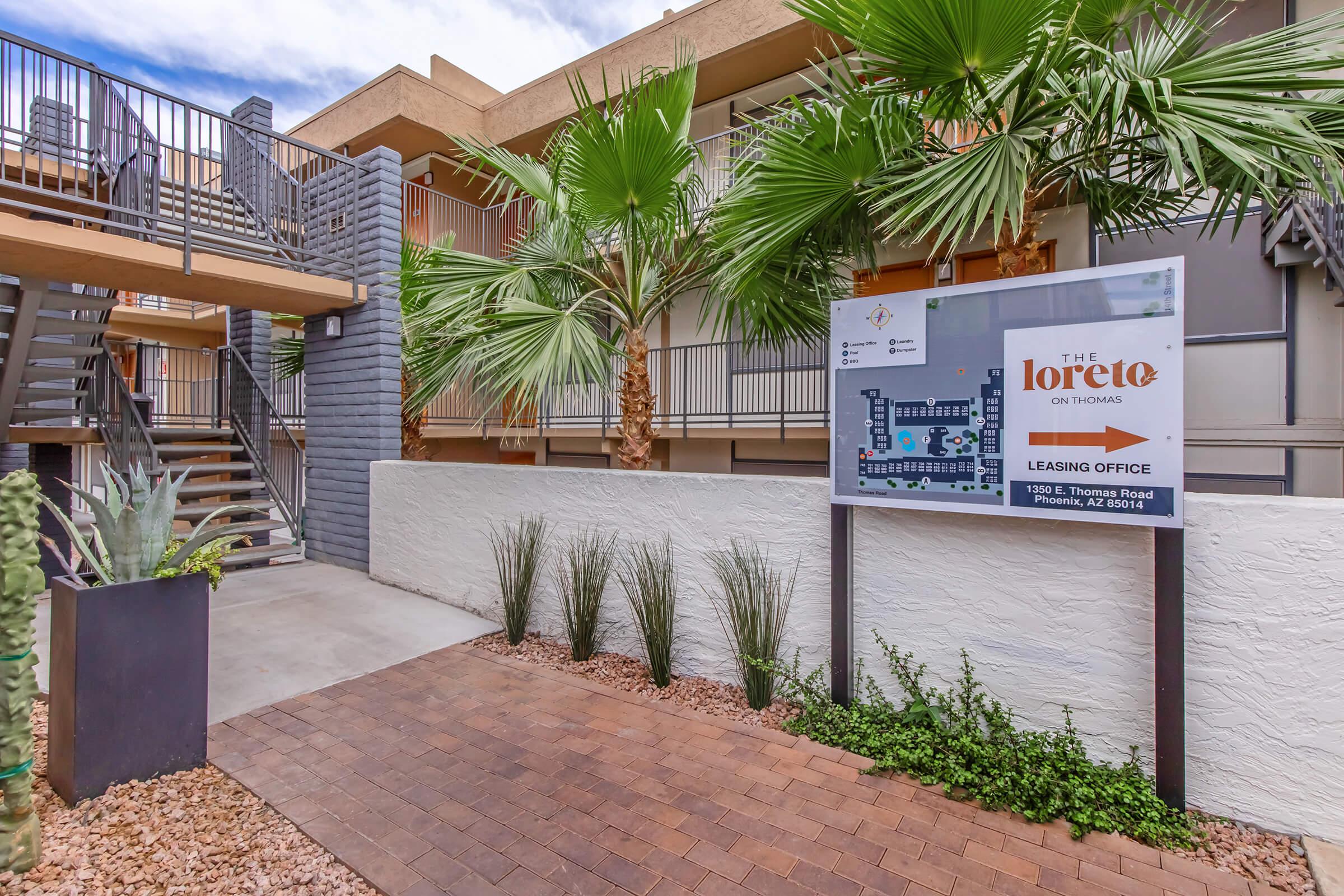A modern apartment complex entrance featuring a leasing office sign for "The Loretto on Thomas." There are palm trees and decorative plants lining a brick pathway leading up to the entrance. The building has a staircase and a contemporary aesthetic with a mix of materials.