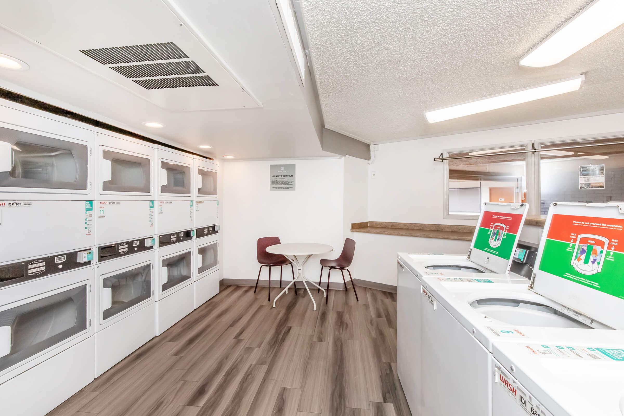A clean and modern laundry room featuring rows of washing machines and dryers. There is a small round table with two chairs for seating. The flooring is a sleek, dark wood design, and the walls are painted white, creating a bright and inviting atmosphere for laundry tasks.