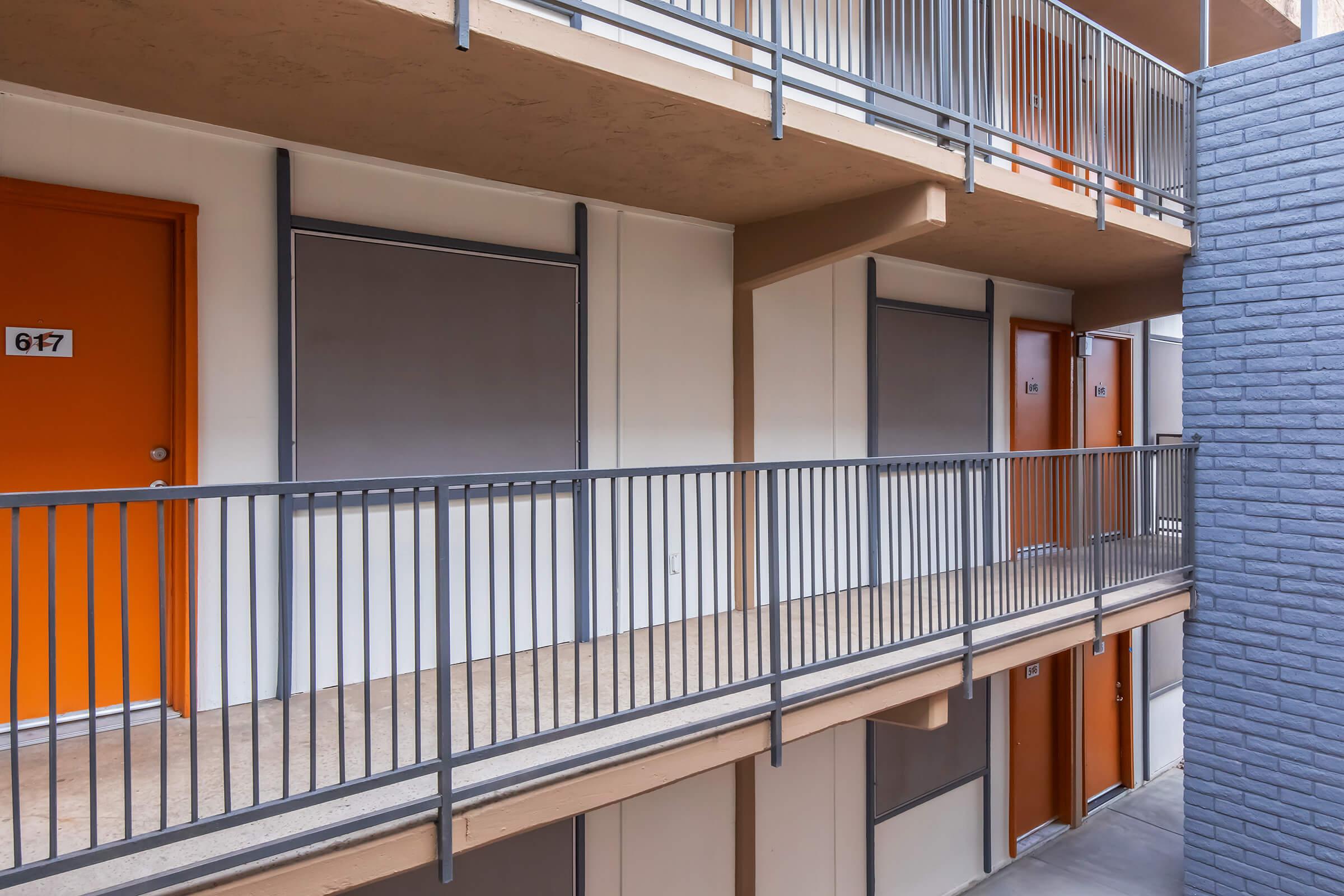 A view of a multi-story apartment building showcasing several balcony levels with orange doors. The balconies have metal railings, and some windows are covered. The building features a mix of light and dark exterior walls, with the adjoining wall in a darker shade.