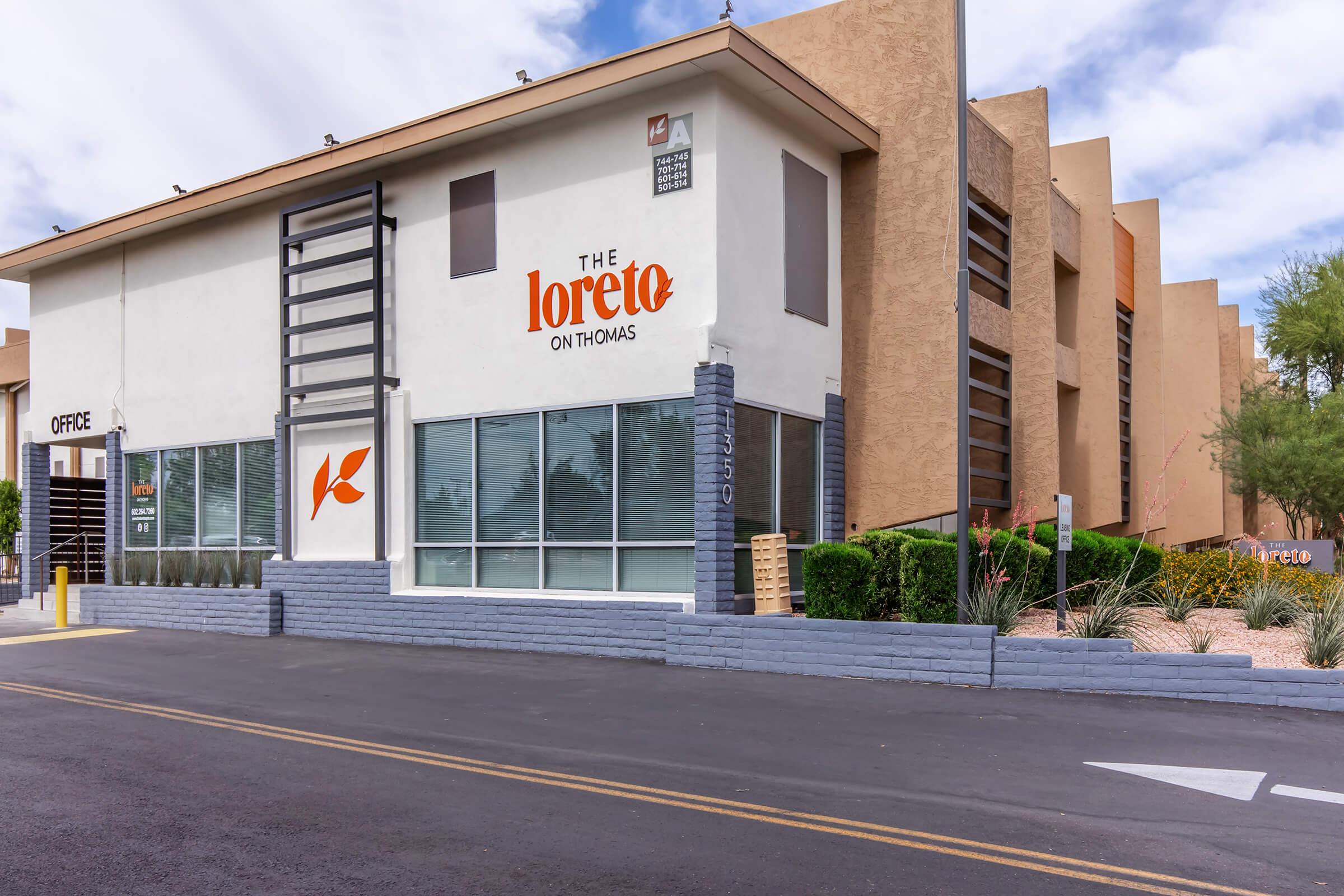 A modern apartment building named "The Loreto on Thomas," featuring large windows and an office area. The exterior has a mix of beige and gray colors, with landscaping visible. A large sign with the name is prominently displayed. The street in front is lined with palm trees and desert plants.