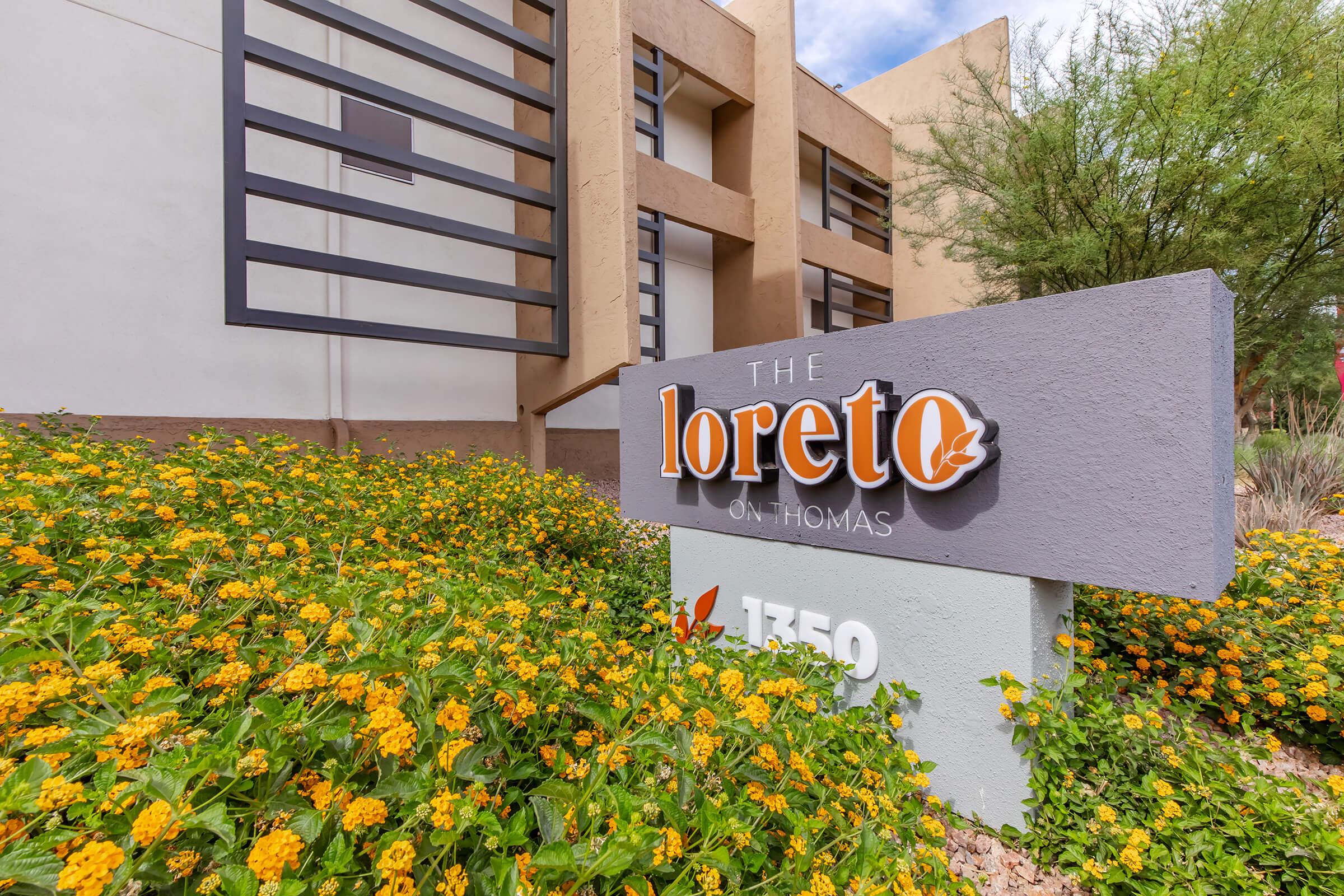 Sign for "The Loreto on Thomas" at 1350, surrounded by vibrant yellow flowers and greenery. The building features modern architectural elements with large windows.