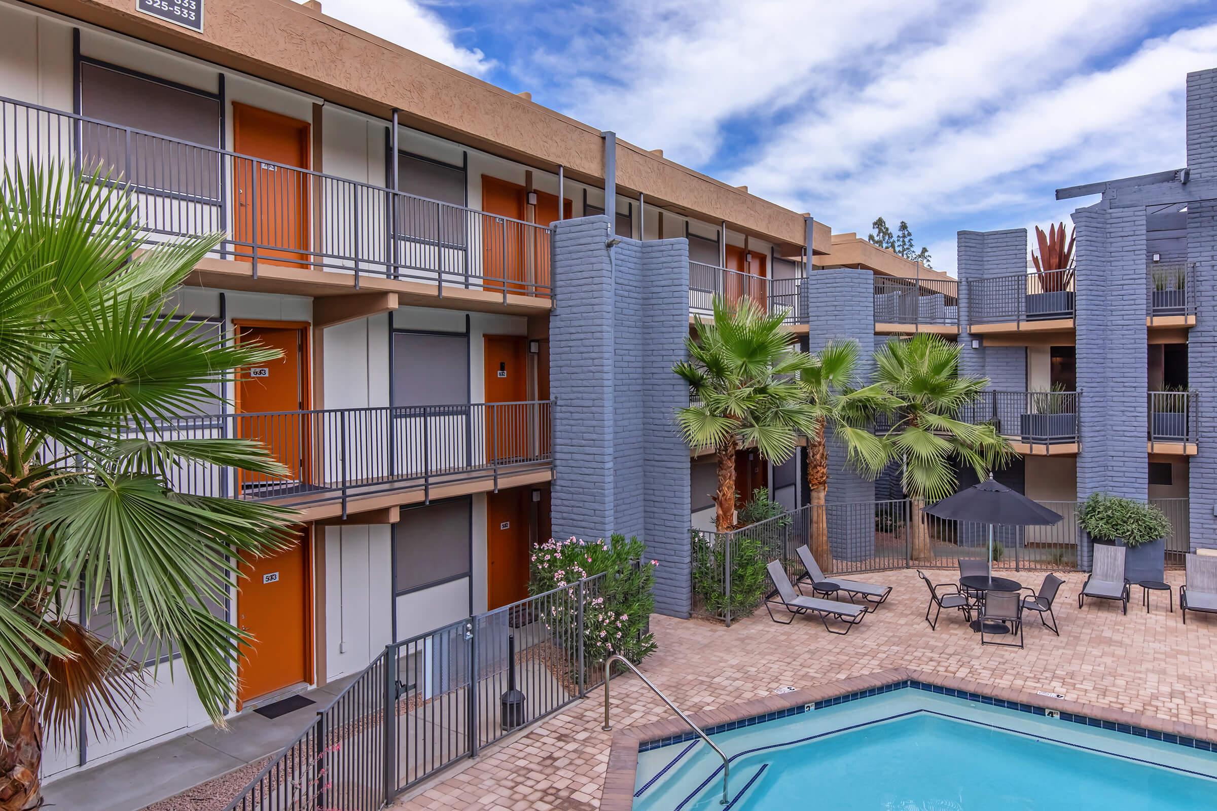 A view of a modern apartment complex featuring a swimming pool surrounded by palm trees and lounge chairs. The two-story building has several orange door entrances and blue-gray exterior walls, set against a backdrop of a partly cloudy sky.