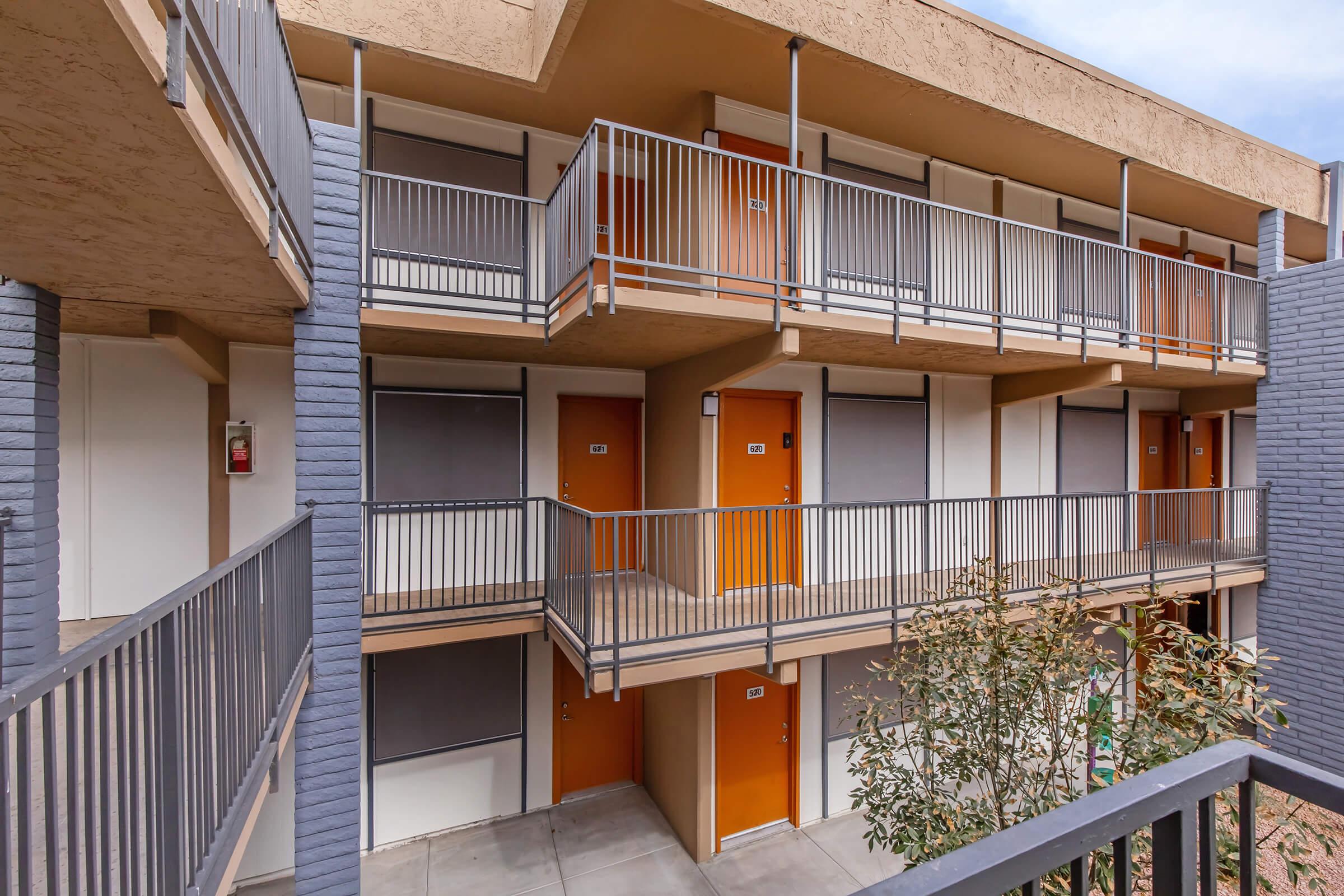 A view of a modern apartment building featuring multiple floors with balconies. The exterior has gray and beige walls, and there are orange doors for the apartments. The area is surrounded by plants, and the balconies have metal railings. The image captures a clean and contemporary architectural design.