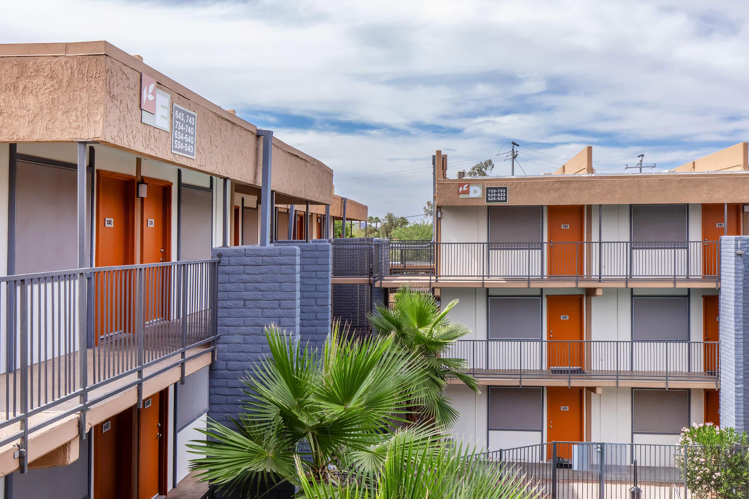 A view of a multi-story motel with orange doors and gray walls. The design features balconies and palm trees in the foreground, under a partly cloudy sky. The building is arranged in a square layout with multiple entrances visible.