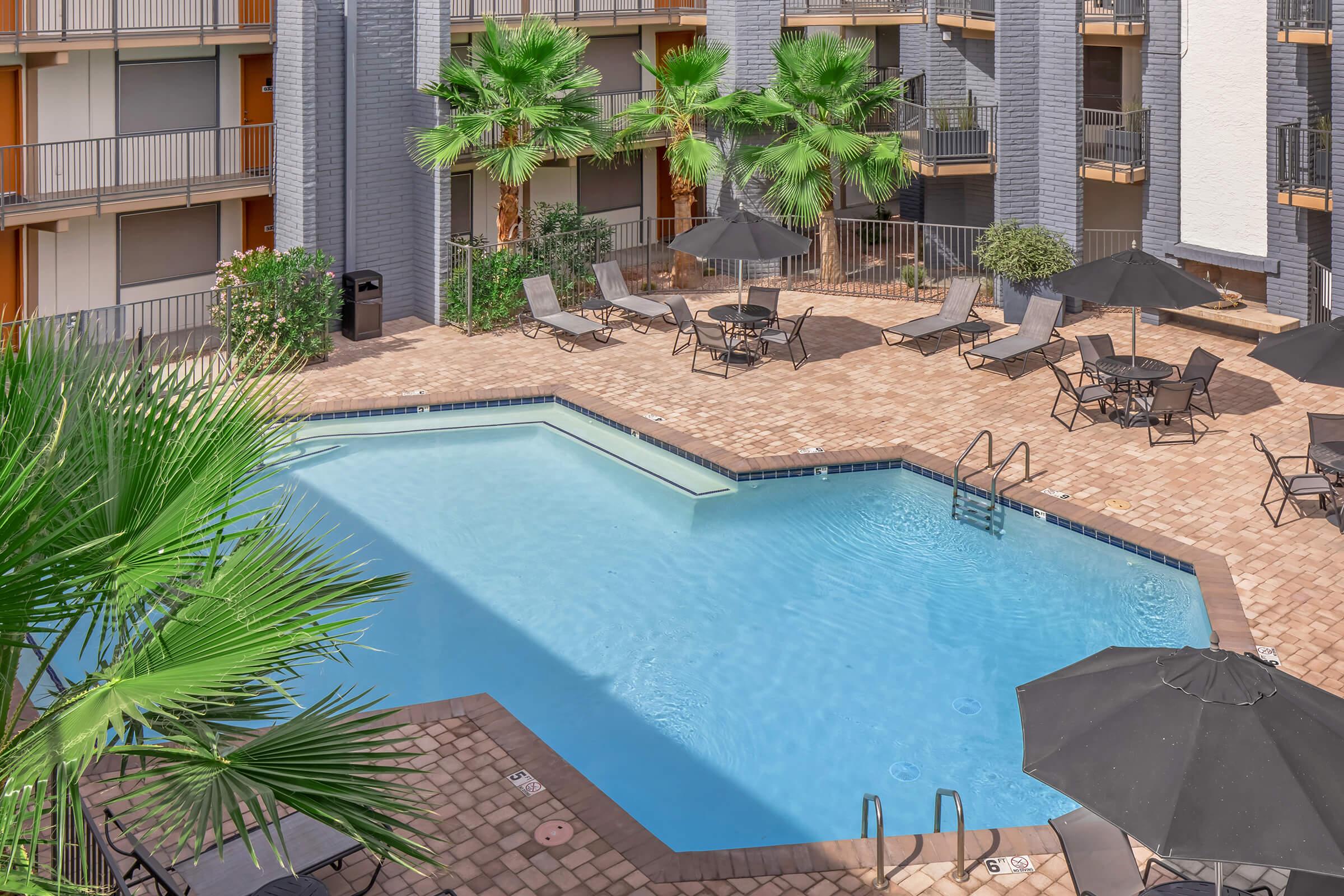 A clear blue swimming pool surrounded by lounge chairs and umbrellas, with palm trees and landscaped greenery in a courtyard setting. The pool area features a brick patio and is framed by nearby apartment buildings.