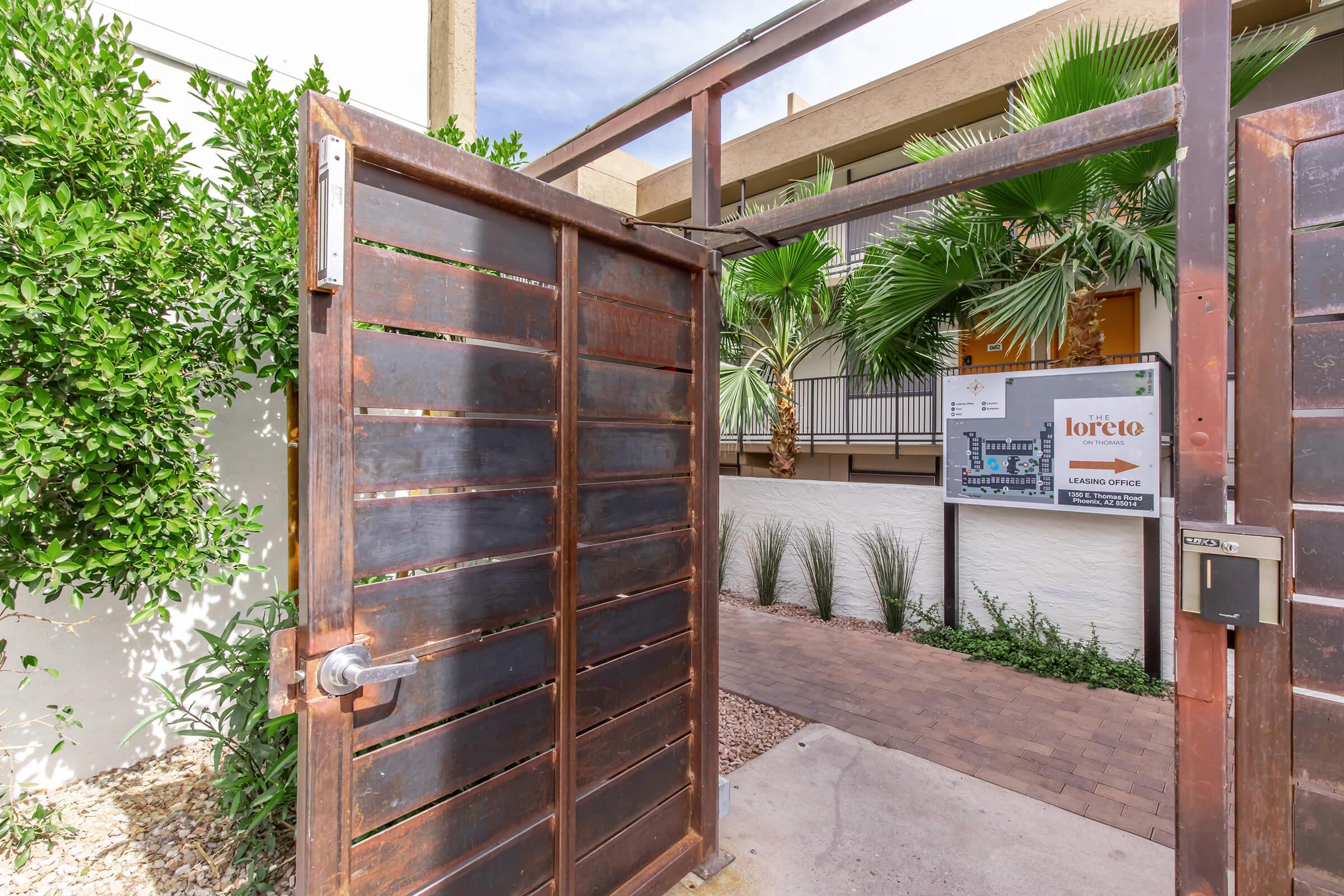A metal gate opens to a pathway leading through a landscaped entrance. The area features palm trees and shrubs, with a sign on the wall indicating the location as "Loreto Learning Office." The pathway is paved and bordered by decorative gravel, leading to a building in the background.
