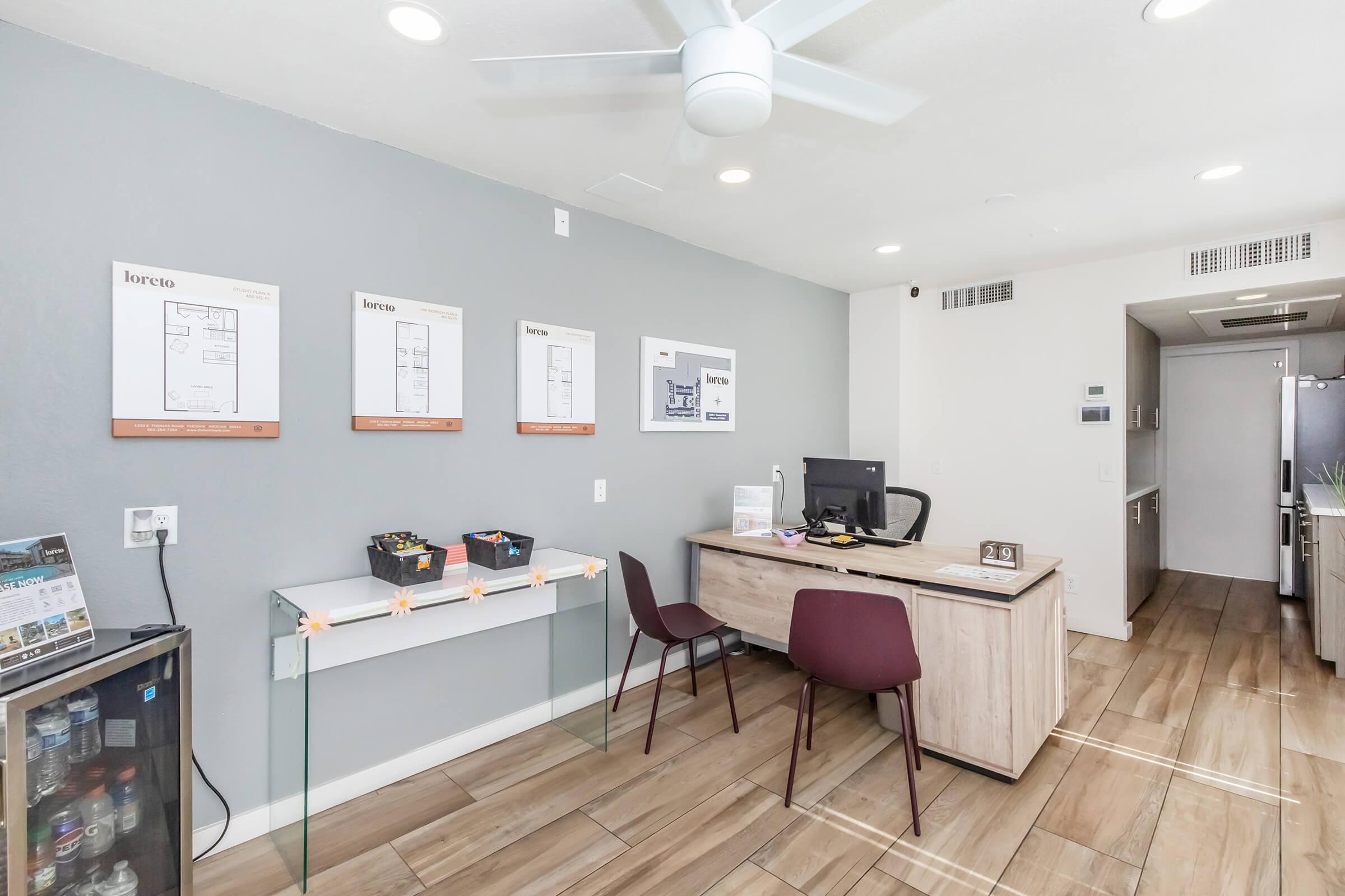 A modern office space featuring a reception area with a desk, a computer, and two chairs. The wall displays four framed architectural plans, and there is a glass table with brochures. The flooring is wooden, and a kitchen area is visible in the background. A ceiling fan is also present.