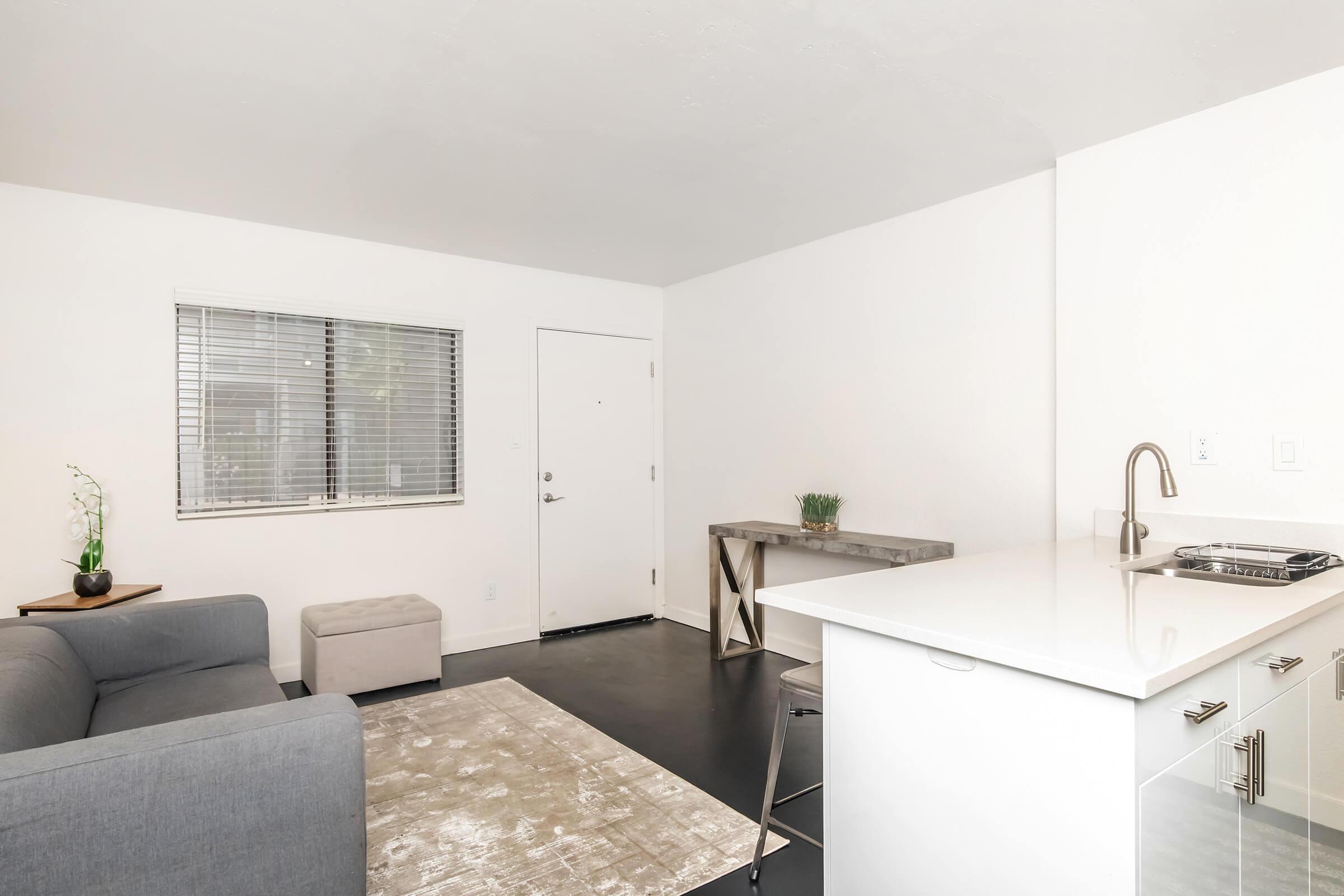 A bright and modern living space featuring a gray couch, a small ottoman, and a minimalist table. There is a kitchen area with a sink, and a door leading outside. Natural light streams in through a window with blinds, creating an inviting atmosphere. The floor is dark, contrasting with the light walls.