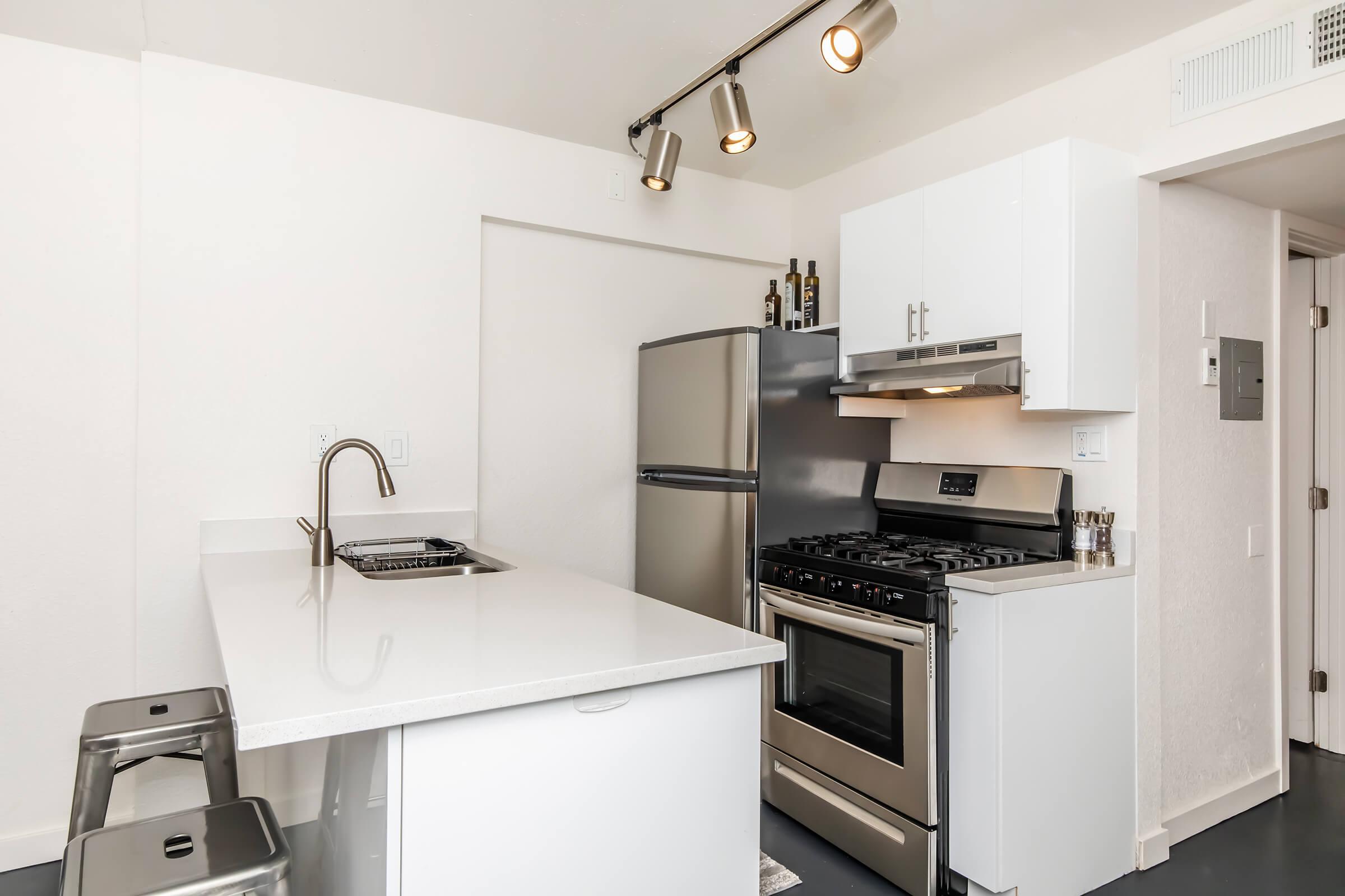 Modern kitchen with a white countertop, stainless steel appliances including a refrigerator and a gas stove, and sleek metal bar stools. The walls are painted white, and there are track lights above the kitchen area. A clean, minimalist design emphasizes functionality and style.