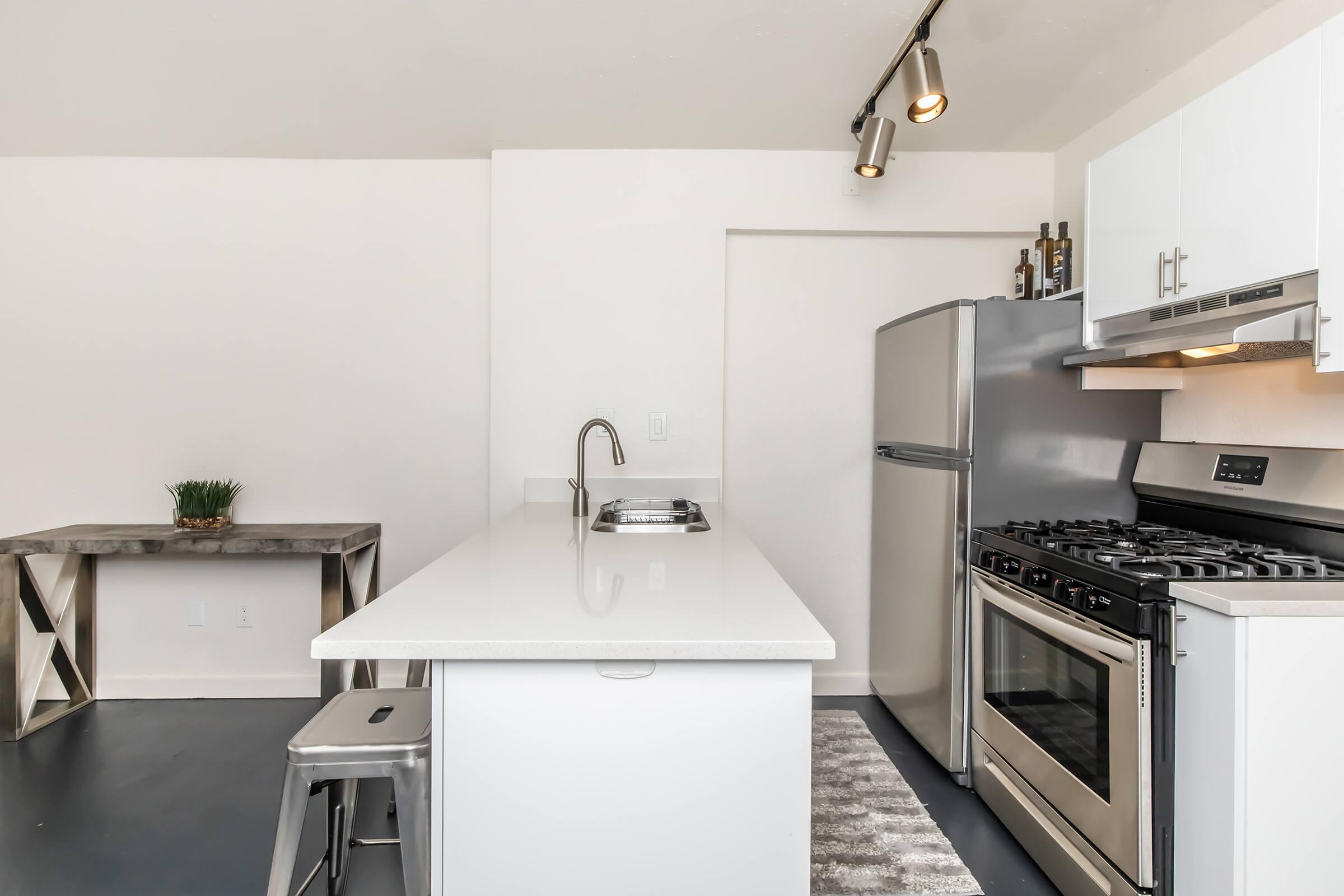 Modern kitchen featuring a minimalist design with white cabinets, a stainless steel refrigerator, and a gas stove. A sleek countertop with a sink is complemented by a small table in the background. The floor is dark, adding contrast to the light walls and fixtures, creating a contemporary feel.