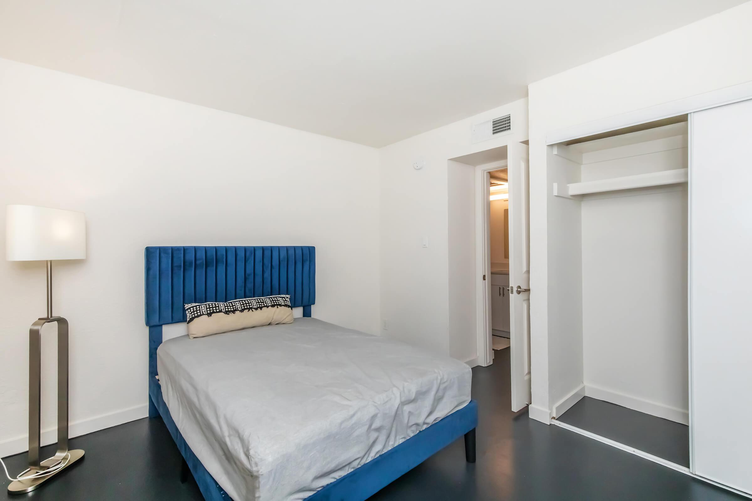 A simple bedroom featuring a blue upholstered bed with a gray mattress and a decorative pillow. There is a modern floor lamp beside the bed, and a closet with sliding doors is visible on the right. The walls are painted white, and the flooring is dark. A bathroom door is partially seen in the background.