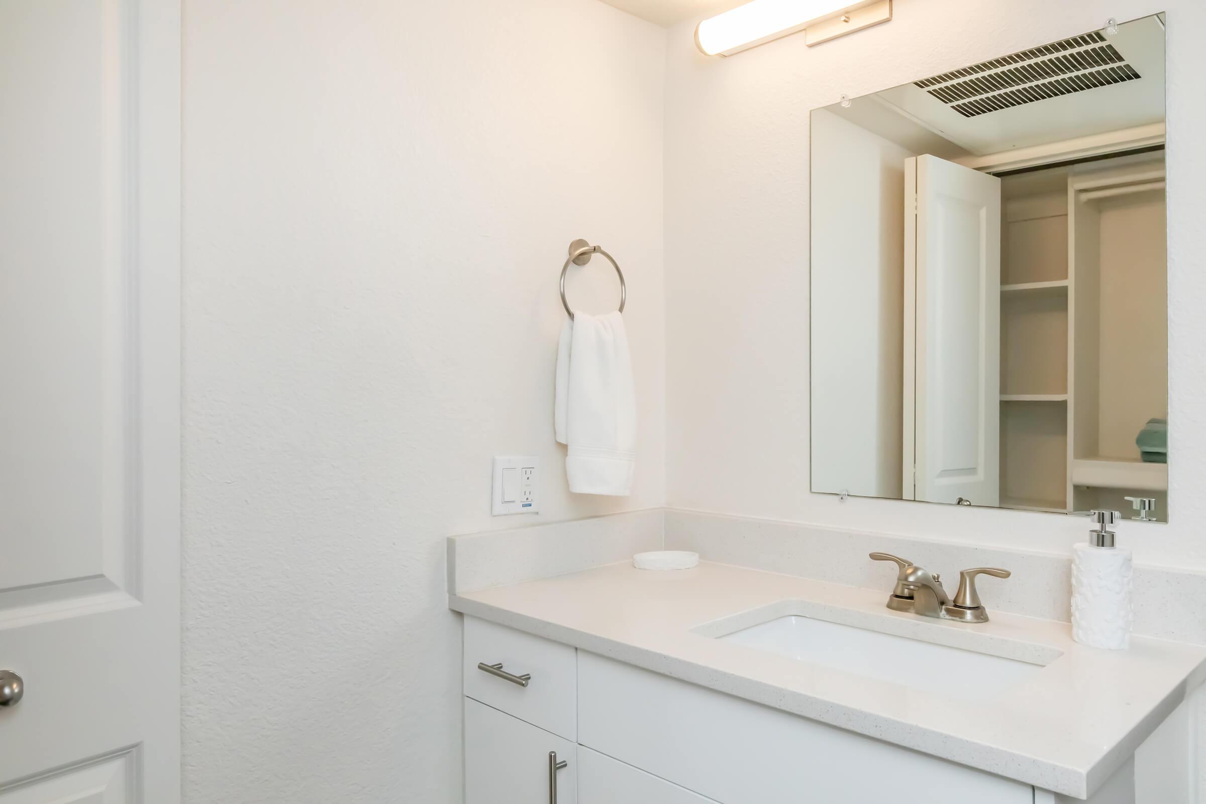 A modern bathroom with a white countertop sink, faucet, and mirror. A towel is hanging on a ring next to the sink. The walls are light-colored, and a cabinet is visible in the background with open shelves. Soft lighting illuminates the space, creating a clean and airy feel.