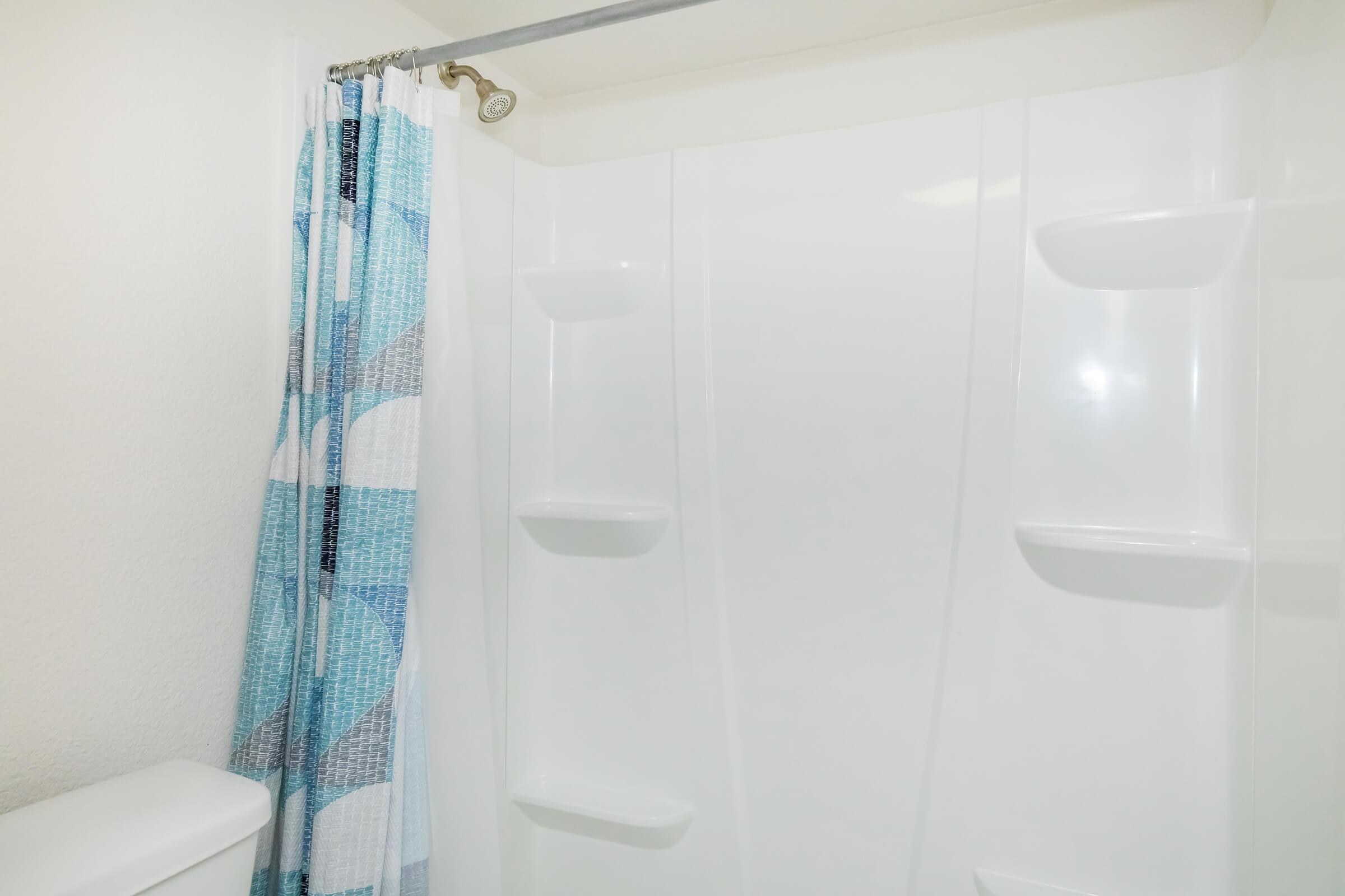 A clean and modern shower space featuring a white shower wall, a blue and white patterned shower curtain on a curved rod, and built-in shelves for toiletries. The area is well-lit with neutral-colored walls.