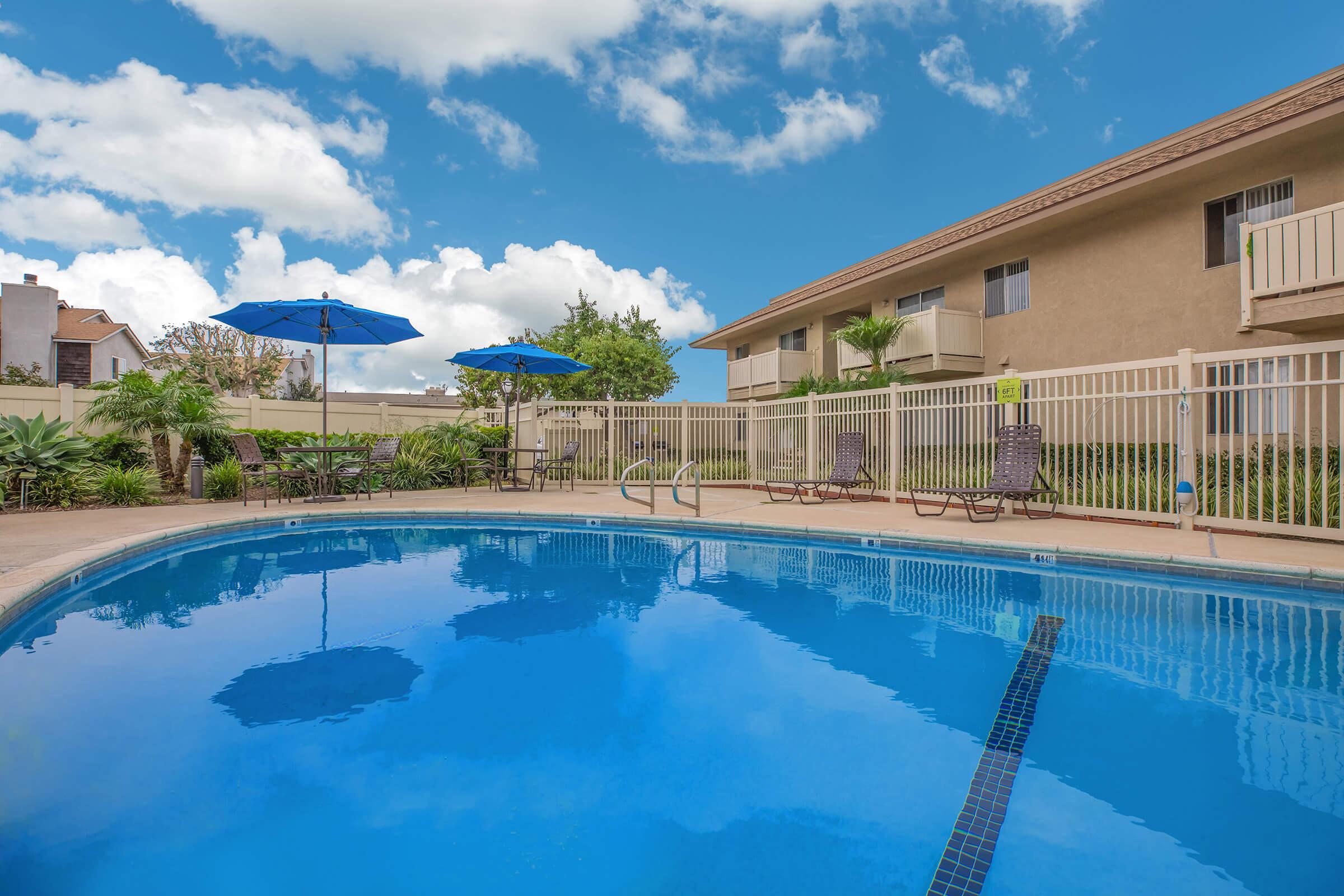 A clear blue swimming pool surrounded by lounge chairs and umbrellas, with a fence in the background. Lush greenery and a residential building are visible, under a bright sky with fluffy white clouds. The inviting scene suggests a relaxing outdoor space.