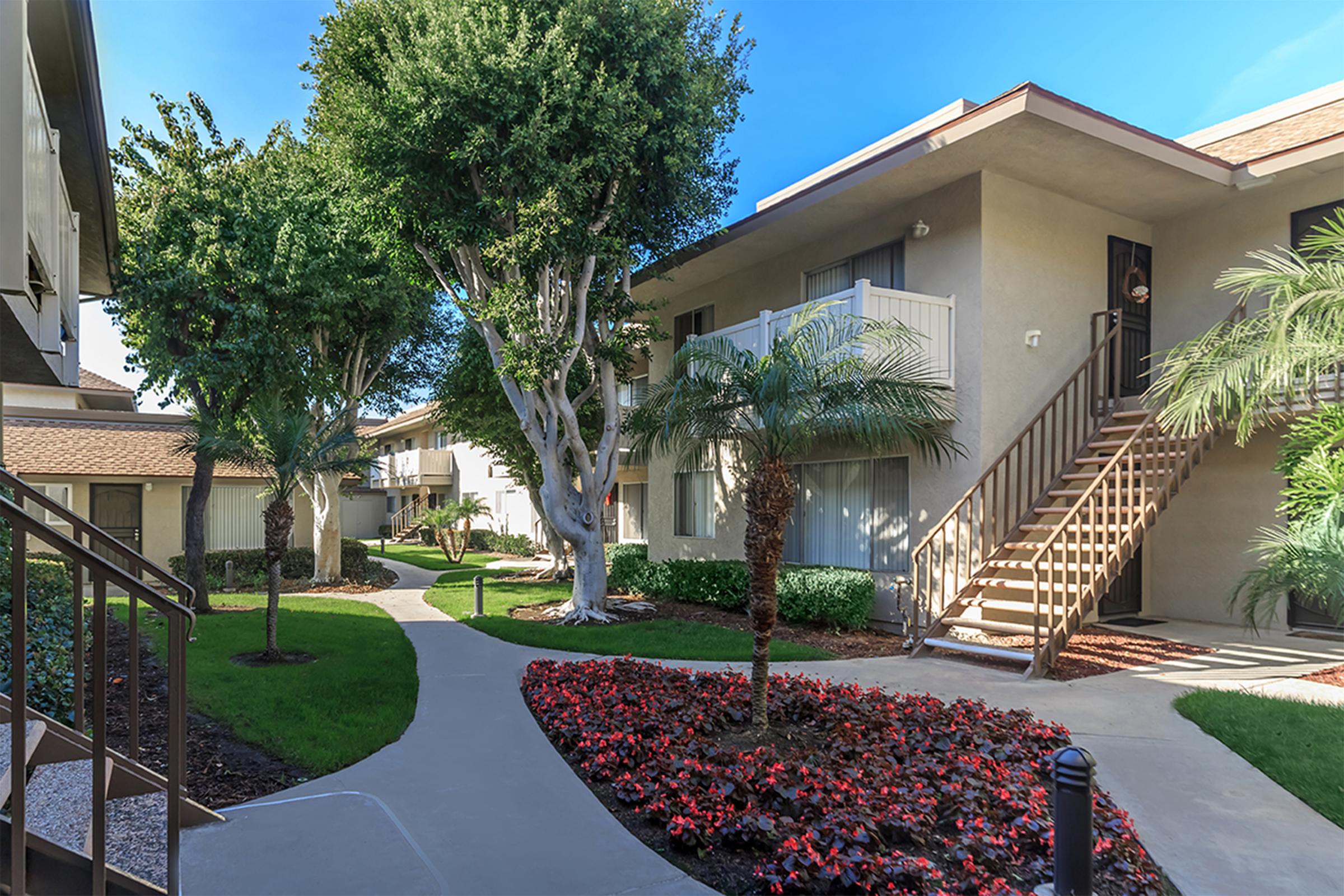 A landscaped courtyard with lush greenery, including palm trees and flowering plants. Two apartment buildings are visible, with balconies and stairs leading to entrances. A winding pathway connects the elements, and the scene is bright and well-maintained under a clear blue sky.