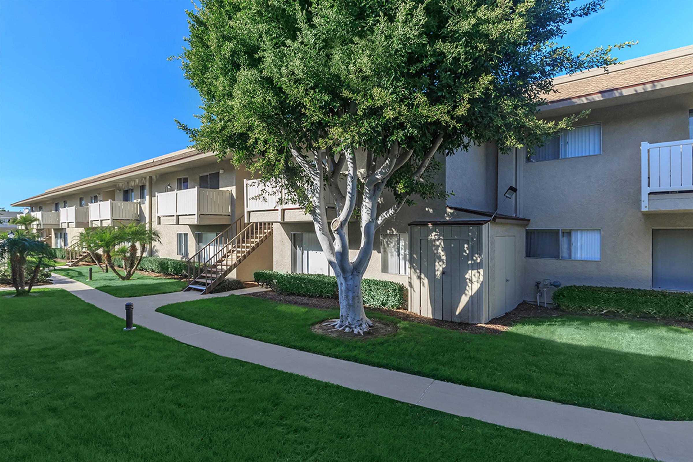 A well-maintained apartment complex featuring two-story buildings with balconies, surrounded by grassy lawns and neatly trimmed bushes. A tree stands in front of the building, and a concrete pathway leads through the landscaped area, offering a clear view of the peaceful outdoor setting.